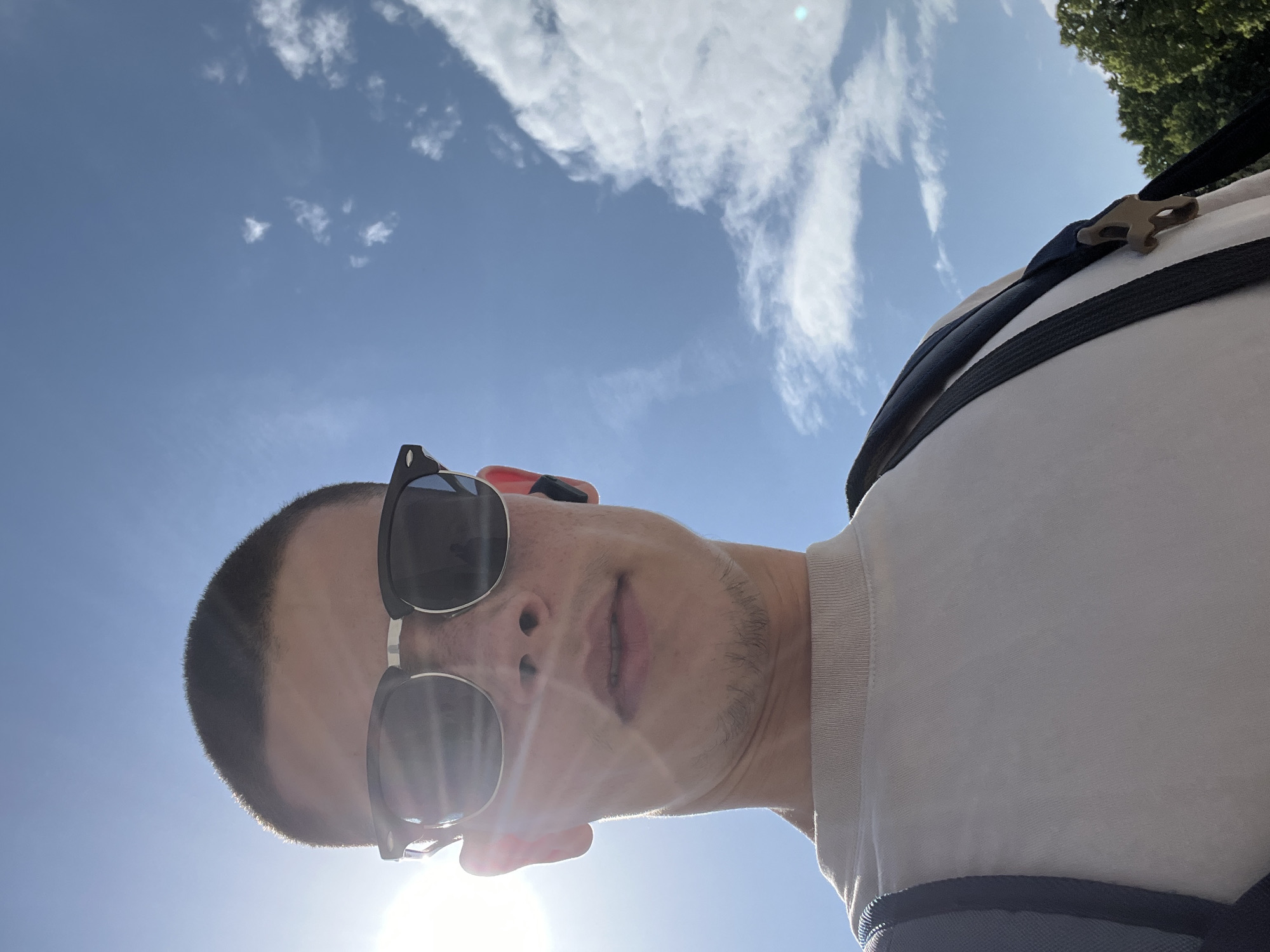 A low-angle selfie depicts a man with a short haircut and stubble, wearing dark sunglasses. He is positioned against a bright blue sky scattered with white clouds. The sun, situated at the bottom of the frame, creates a lens flare. The man is wearing a light-colored T-shirt and a backpack with dark straps and a buckle detail visible on one shoulder. The location is not discernible as Plovdiv, Bulgaria from the given image.