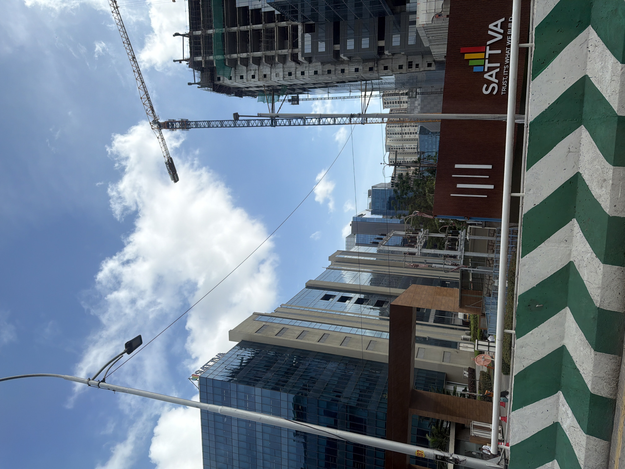 This image shows a construction site in Hyderabad, India, with several modern office buildings. A tall tower crane is visible, actively engaged in construction work on a high-rise building. In the foreground, there is a concrete barrier with green and white chevron markings. A large sign with the logo "SATTVA" and the tagline "TRUST, IT'S WHAT WE BUILD" is prominently displayed, indicating the developer or project name. The sky is blue with scattered white clouds, suggesting it's daytime. The overall scene depicts urban development and infrastructure growth.