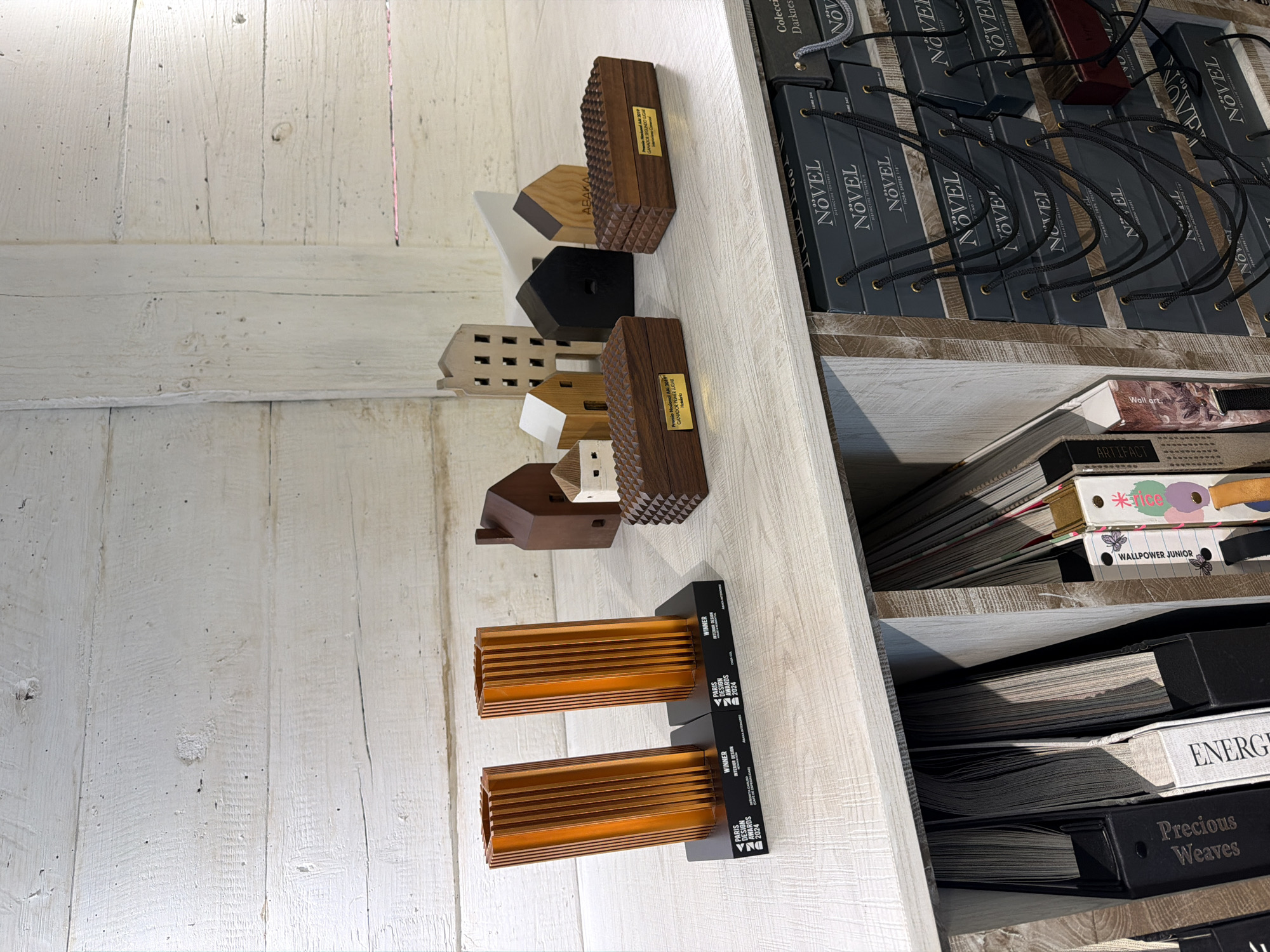 The image depicts a shelf containing various items. On the top shelf, there are several grey boxes labeled "NÖVEL" with black cords attached. The shelf below holds a stack of books or catalogs with visible titles like "ARTIFACT," "WALLPOWER JUNIOR," "jice," "ENERG," and "Precious Weaves." On the surface in front of the shelf, there are two awards labeled "PARIS DESIGN AWARDS 2024" and several small, stylized wooden building models. One of these wooden models has an inscription on its side, "Premio Nacional AAI 2019, GANADOR PRIMER LUGAR Hotelero." Another wooden model displays "ABAKA". The background consists of a whitewashed wooden wall. The location is inferred to be in Puebla, Mexico, based on the Spanish inscription "Premio Nacional AAI 2019" and the label "ABAKA," a brand with a presence in Mexico.