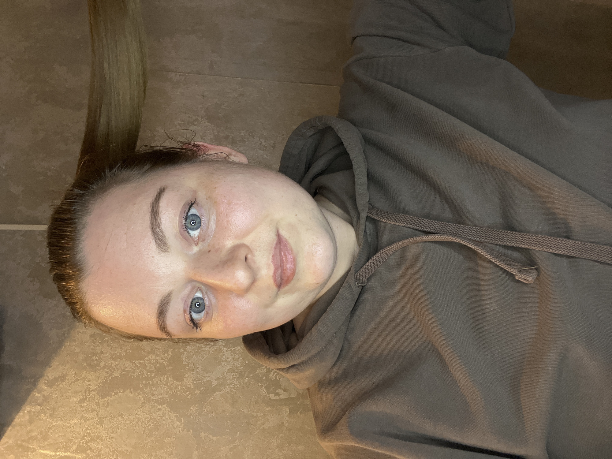 A woman is lying on a beige tile floor, wearing a brown hooded sweatshirt. She has fair skin, blue eyes, and her blonde hair is pulled back. The location is unknown and not determined by the image.