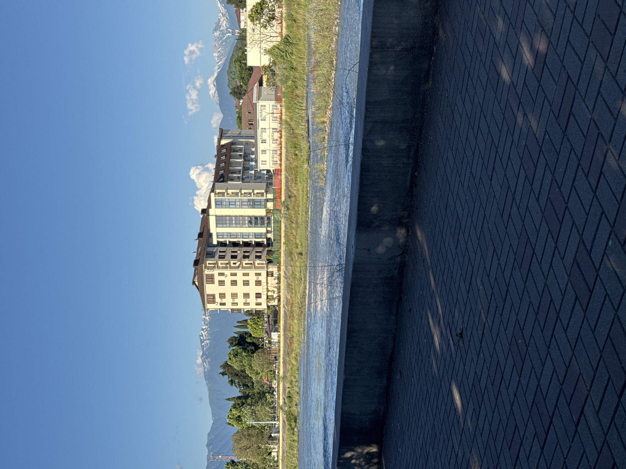 The image depicts a wide, fast-flowing river with a concrete embankment in the foreground. Beyond the river, a multi-story building with a light-colored facade and balconies is visible, set against a backdrop of lush green trees and snow-capped mountains. The sky is a clear, bright blue with a few scattered clouds. The pavement in the foreground is laid with rectangular pavers arranged in a herringbone pattern, with shadows cast by an unseen light source. The overall scene suggests a tranquil, scenic location within a city or resort area. It is plausible that this location is in Sochi, Russia, given the architectural style of the building and the mountainous terrain visible in the background, characteristic of the region.