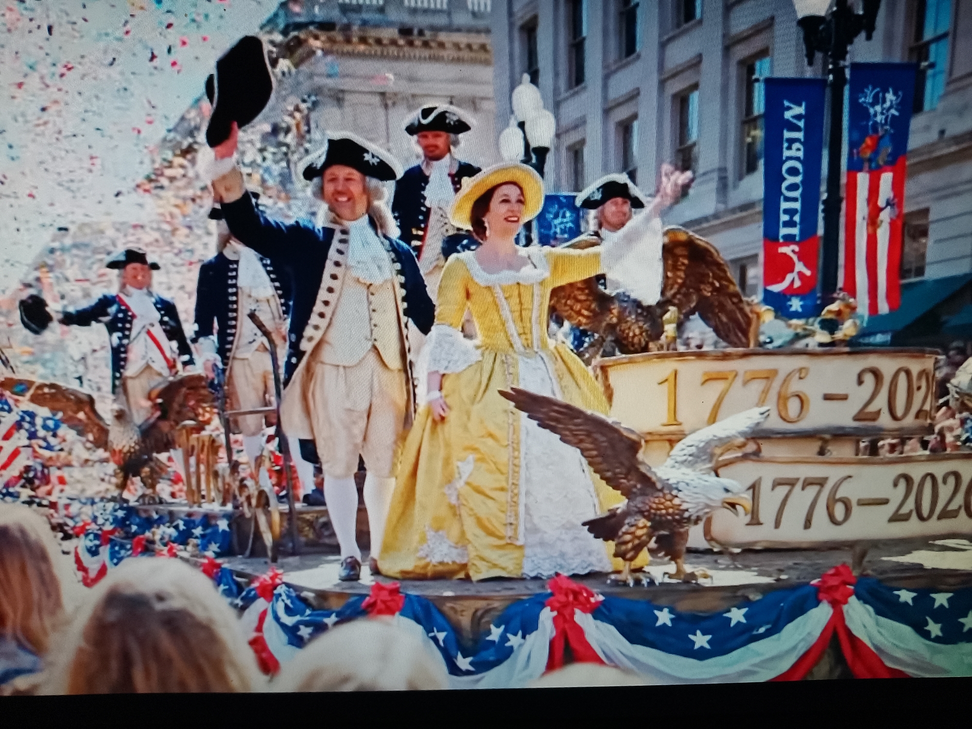 The image depicts a parade scene with people dressed in colonial-era attire, celebrating an event that seems to commemorate the year 1776. Several individuals in tricorn hats and period clothing are visible, some waving and throwing confetti. One woman in a yellow gown stands out. The parade features floats adorned with eagles, American flags, and banners with the text "1776-2020". The backdrop appears to be an urban setting with buildings. The reference to Chiajna, Romania, in the prompt is not supported by any visual cues within the image itself.