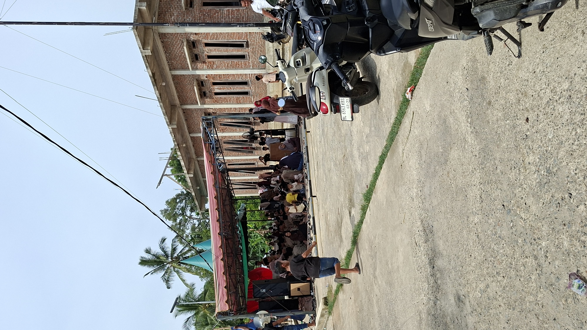 The image depicts an outdoor gathering in Asan, Indonesia, likely a community event or religious service, based on the presence of people seated under a covered area and a building in the background that resembles a mosque or community hall. A group of people, including men, women, and children, are seated on the ground under a red-and-white striped canopy supported by metal poles. Some individuals appear to be listening to a speaker, as evidenced by the presence of large speakers on the left side of the canopy. A young boy in a black t-shirt and denim shorts is running in the foreground, facing away from the camera. In the background, a brick building with arched windows and a concrete roof is visible. Several motorcycles are parked in the foreground, suggesting the mode of transportation for attendees. The environment is paved, with a strip of grass running along the edge of the paved area. The sky is clear and blue, indicating a daytime scene. The overall impression is one of a modest community gathering in a semi-urban or rural setting.
