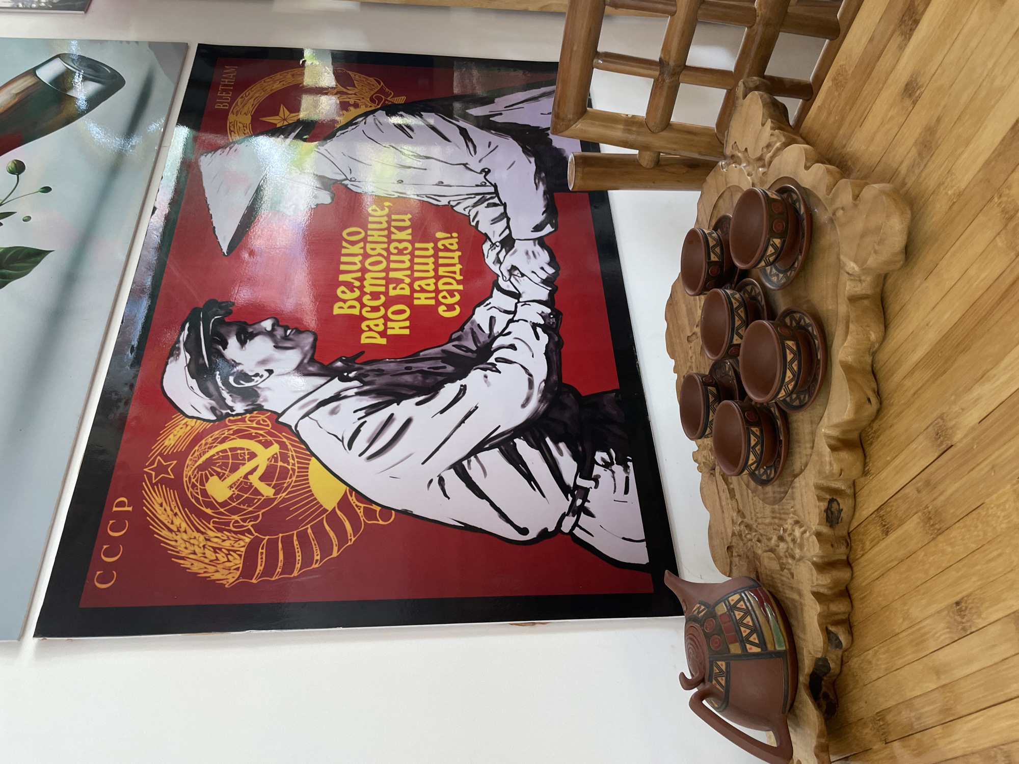 A red and black framed poster titled "VIETNAM" and labeled with "CCCP" and featuring two stylized figures clasping hands is mounted on a white wall. Adjacent to the poster is a wooden, decorative display rack holding six small brown teacups and saucers, along with a brown teapot with geometric patterns. The rack is mounted on a bamboo wall. The objects are located in Phu Thuy Ward, Vietnam.