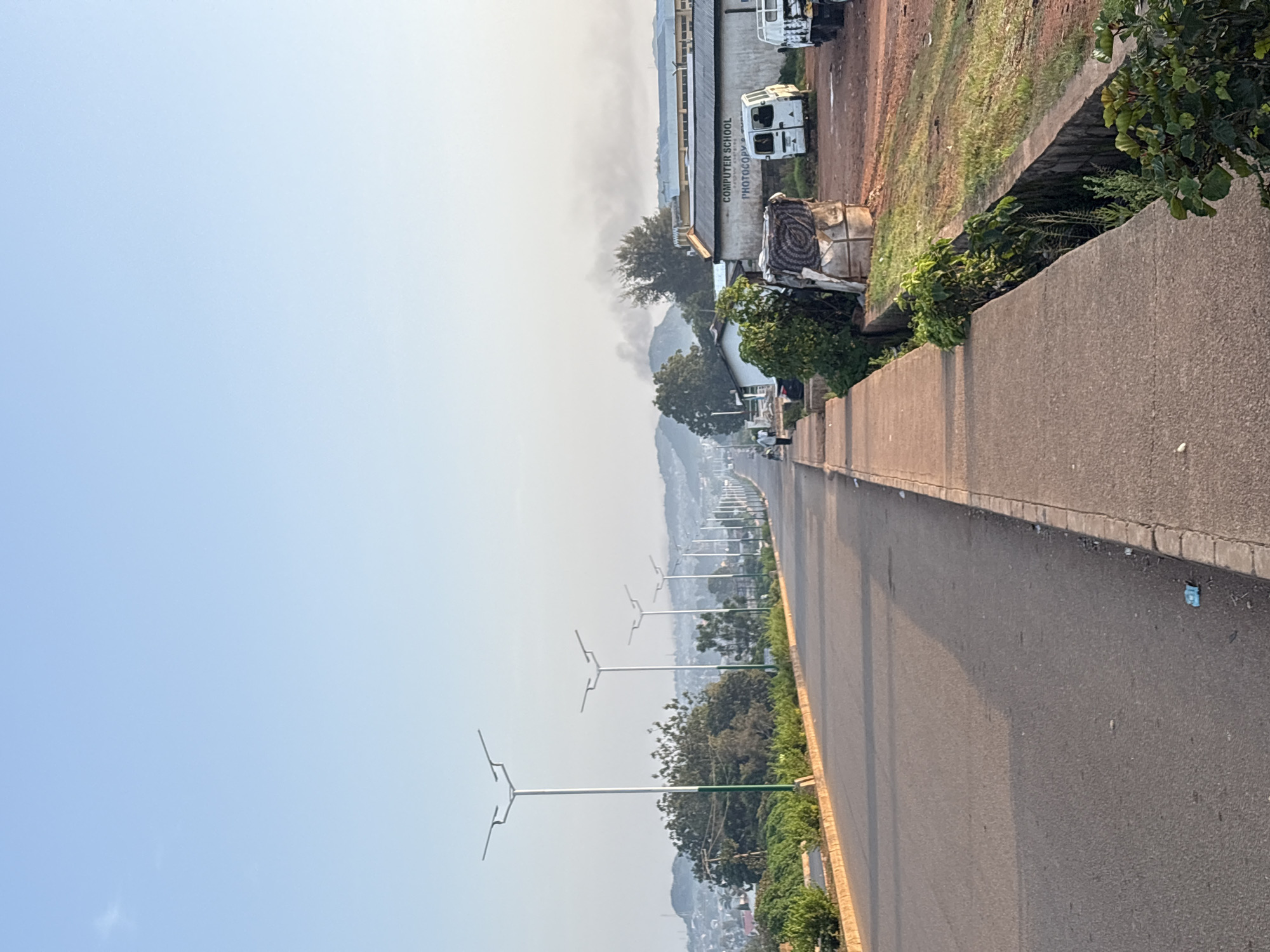 The image depicts a wide asphalt road in Bukuru, Nigeria, lined with streetlights and a concrete sidewalk. The road curves gently towards a hazy horizon dotted with rolling hills and buildings. On the right side of the road, a building with the sign "COMPUTER SCHOOL PHOTOCOPY" is visible, along with parked white vans and a pickup truck. The scene appears to be in the morning, with soft, diffused sunlight illuminating the landscape and casting shadows on the road. Vegetation, including trees and shrubs, is present along the sides of the road and in the foreground. A small trench runs alongside the sidewalk, filled with greenery. A few individuals can be seen walking in the distance on the sidewalk. The overall impression is that of a semi-urban environment in Nigeria.