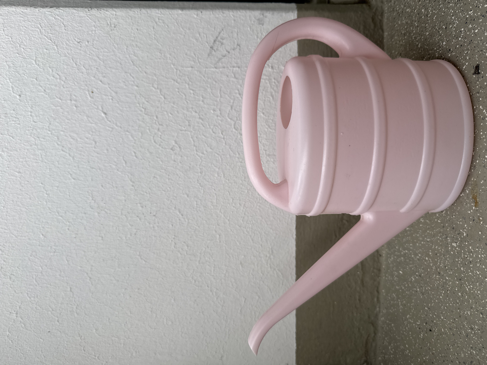 A pink plastic watering can sits on a speckled gray surface, likely a floor or ground. Behind it, a textured white wall extends upwards, with a dark horizontal line forming a border near the bottom of the wall. The watering can features a long spout on the left, a curved handle on the top, and a bulbous body with several horizontal indentations. The lighting suggests it is daytime. The scene appears to be outdoors or in a transitional space, such as a porch or entryway. No people or animals are present in the image.