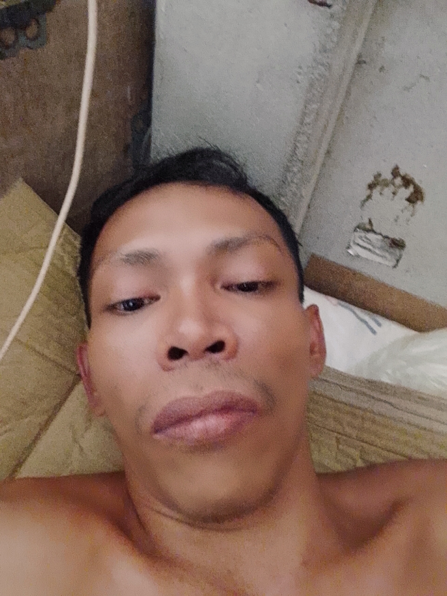 A selfie of a shirtless man with dark hair and brown eyes. He is lying down, and the camera is positioned above his face. Behind him is a white wall with some discoloration and a brown object. There is also a white cable and a brown surface visible. The location is Bukit Ketok, Indonesia. No specific details about the environment are discernible to indicate the exact location within Bukit Ketok.