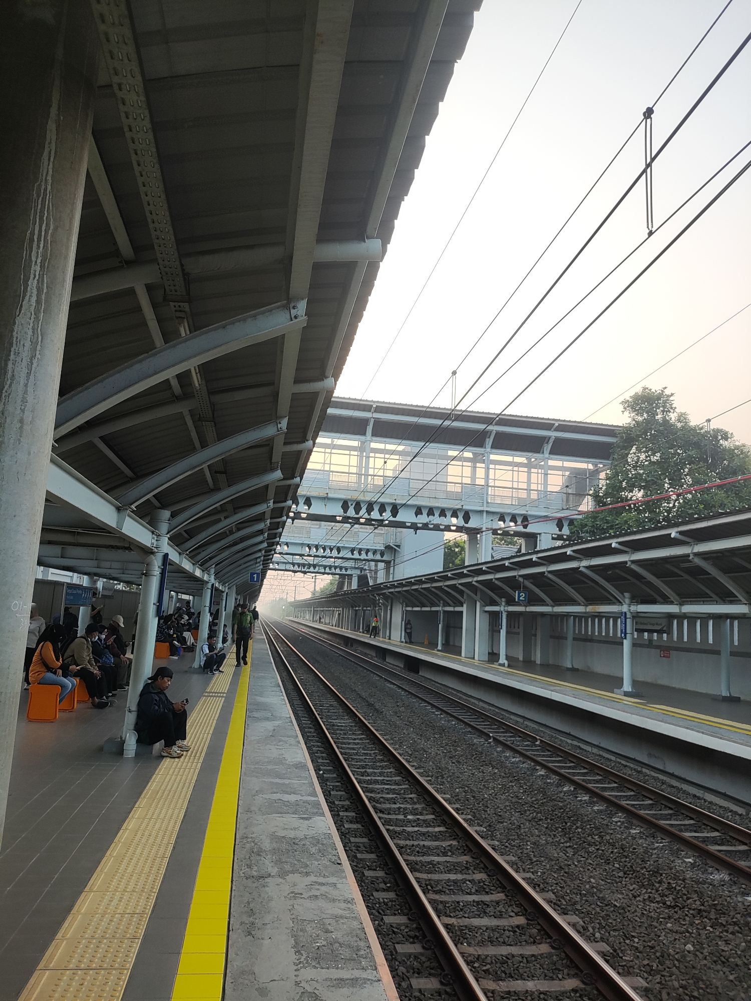 The image depicts a train station platform in Telagamurni, Indonesia, with train tracks stretching into the distance. A covered walkway runs along the platform, supported by metal beams and concrete pillars. Several people are visible on the platform, some sitting on orange benches and others standing or walking. A man in a dark jacket is sitting on the ground near the edge of the platform, looking at his phone. The sky is overcast, with a hazy glow suggesting either early morning or late afternoon. Overhead electrical wires for the trains are clearly visible. The platform has a distinct yellow safety line along the edge. The overall impression is of a functional, albeit somewhat utilitarian, urban transportation hub.
