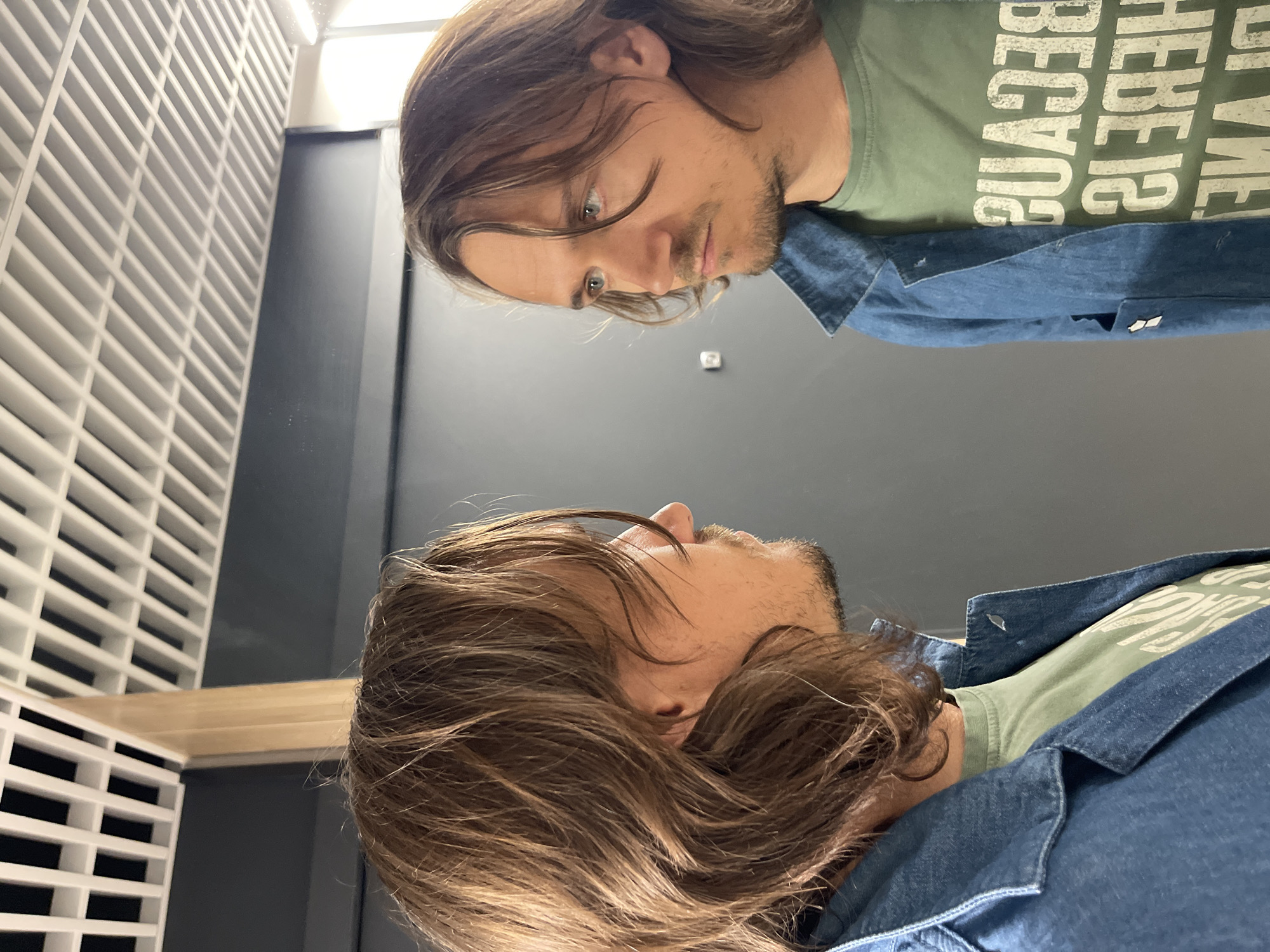 The image depicts a man standing in front of a mirror. The man has light brown hair and is wearing a denim jacket over a green t-shirt. The reflection in the mirror shows the same man, but the text on his t-shirt is reversed. The background consists of a white latticework structure on the left and a dark gray wall. The location is identified as Unknown, Unknown, but specific details about the location within the city are not provided.