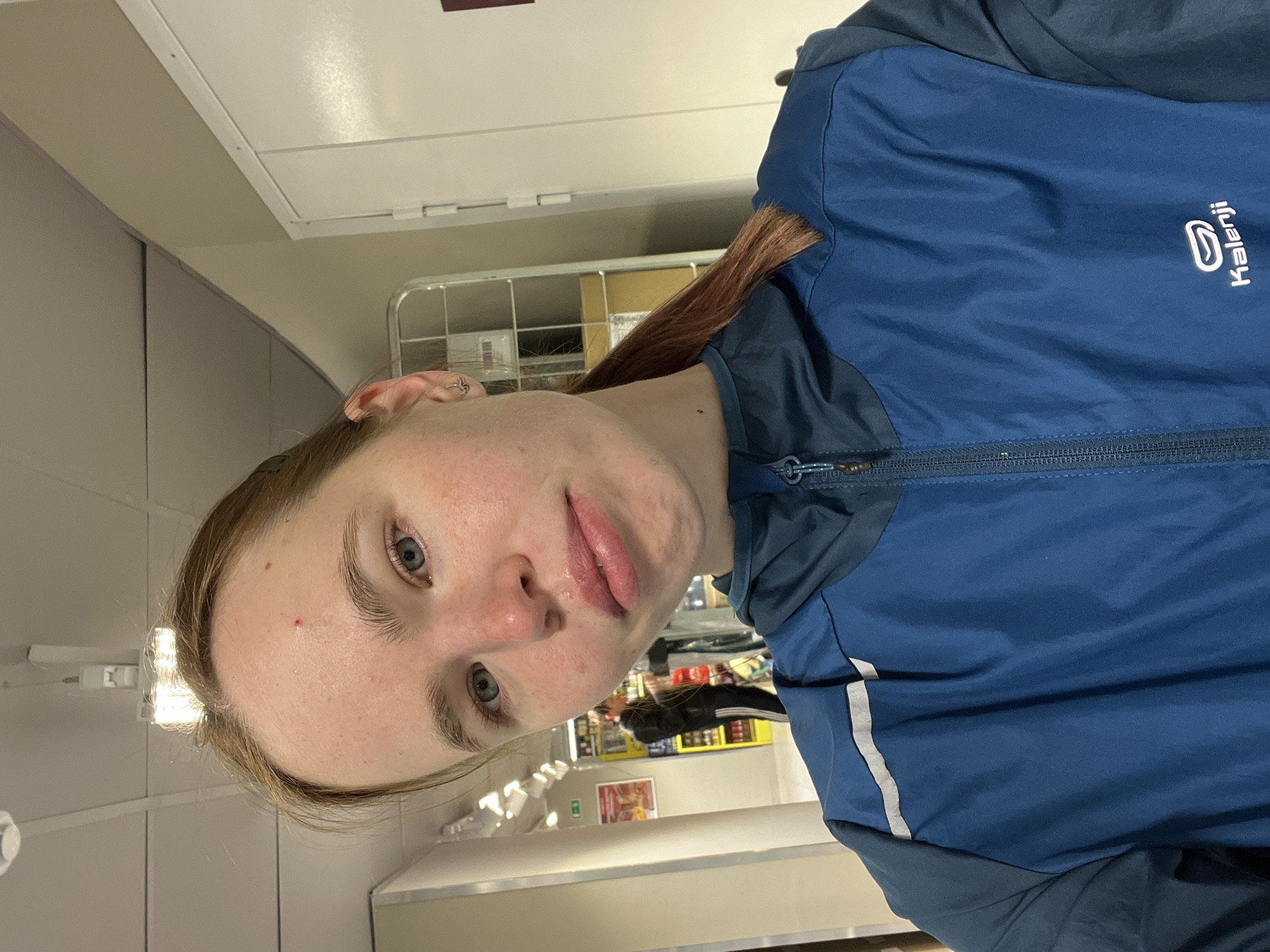 A fair-skinned woman with auburn hair in a ponytail is facing the camera. She has light eyebrows and eyes, and is wearing a dark blue Kalenji jacket. A silver earring is visible in her left ear.
The location appears to be a shop in the city of Perm, Russia. Background details include shelving with various products, likely food items and groceries. A metal cage-like structure sits behind the woman's head. A fluorescent light fixture is mounted on the ceiling.