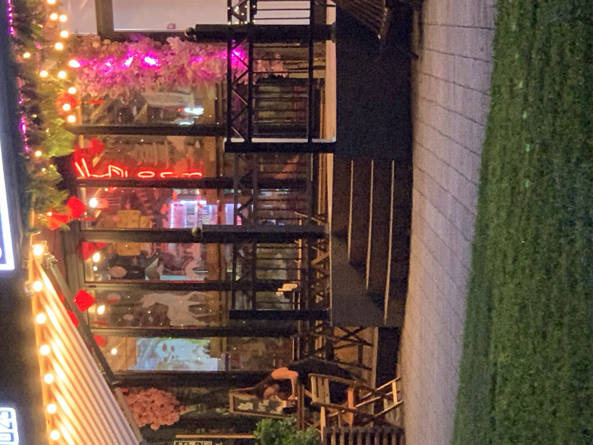 The image shows an outdoor seating area of a restaurant at night. A woman with dark hair and a black top with an orange strap is seated at a table on the left side of the image, looking down at something in front of her. In front of her is a table with two chairs. There is a blackboard to the left of the table. The restaurant has large windows with neon lights inside and is decorated with string lights and pink artificial plants. A set of stairs leads up to the restaurant entrance. A railing separates the seating area from the stairs. The ground in front of the seating area is paved with rectangular stones, and a grassy area is visible in the foreground. The overall lighting is dim, with the string lights and neon signs providing illumination.