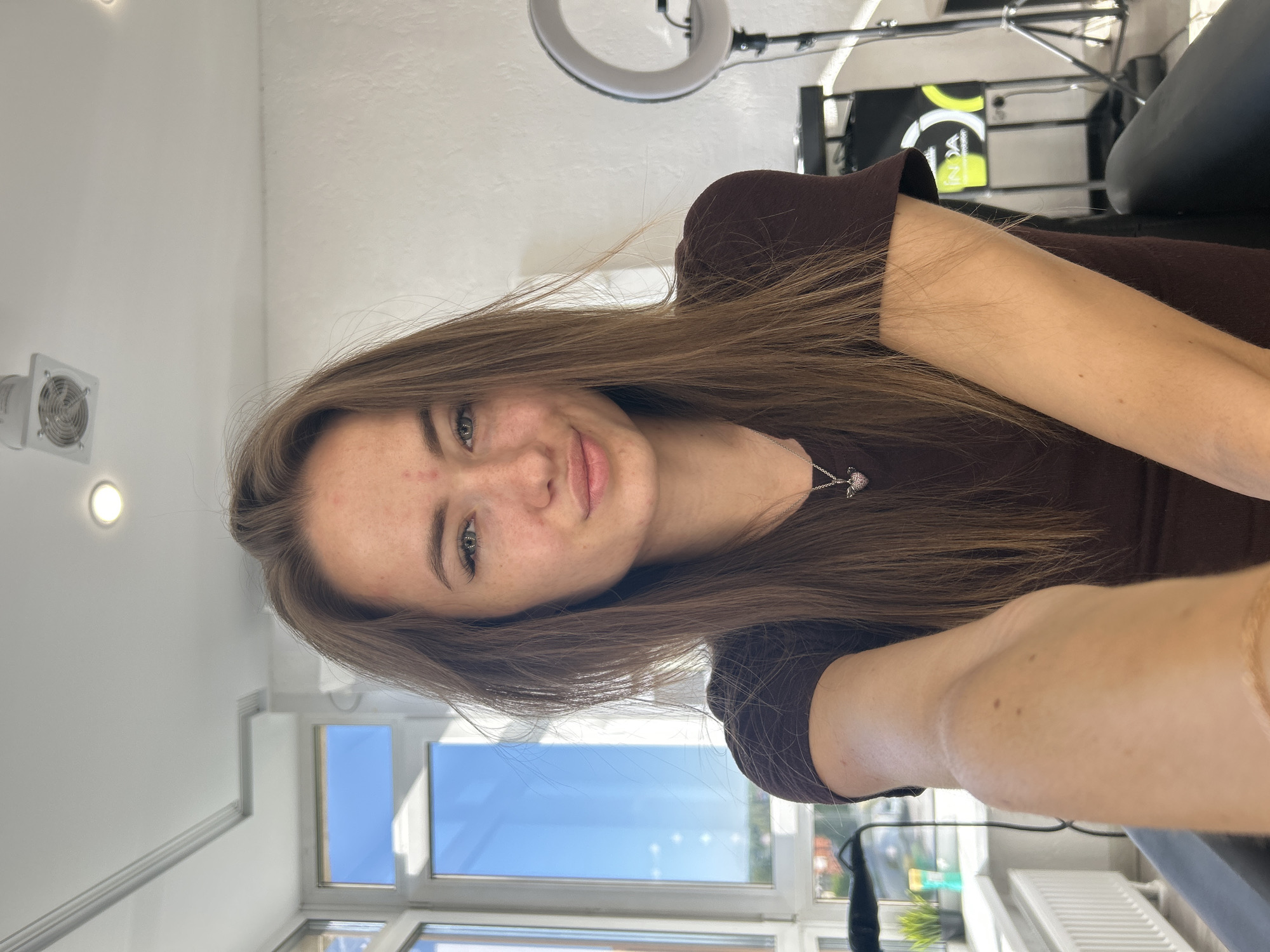 A young woman with long, brown hair, wearing a dark brown t-shirt and a silver necklace with a heart pendant, is captured in a medium shot selfie. She is seated, with her right arm extended towards the camera. Her face is turned slightly towards the lens, and she has a faint smile. The background shows a bright interior space with large windows, a radiator, and professional lighting equipment, suggesting a studio or salon environment. The overall scene is well-lit, with natural light streaming in from the windows.