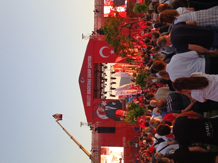 A large crowd of people gathered in front of a stage with a large red banner that reads "Millet İradesine Sahip Çıkıyor" (The Nation Stands Up to Its Will). The stage is adorned with Turkish flags and the image of a man in a suit, likely a political figure. The scene takes place in the Konak district of İzmir, Türkiye. The crowd is dense and comprised of individuals of all ages, dressed in casual clothing. Many are holding Turkish flags, some are taking pictures or videos of the stage and the crowd. There is a large crane to the left of the stage with two individuals working on the crane. The scene is likely a political rally or public event, held outdoors under a clear sky.