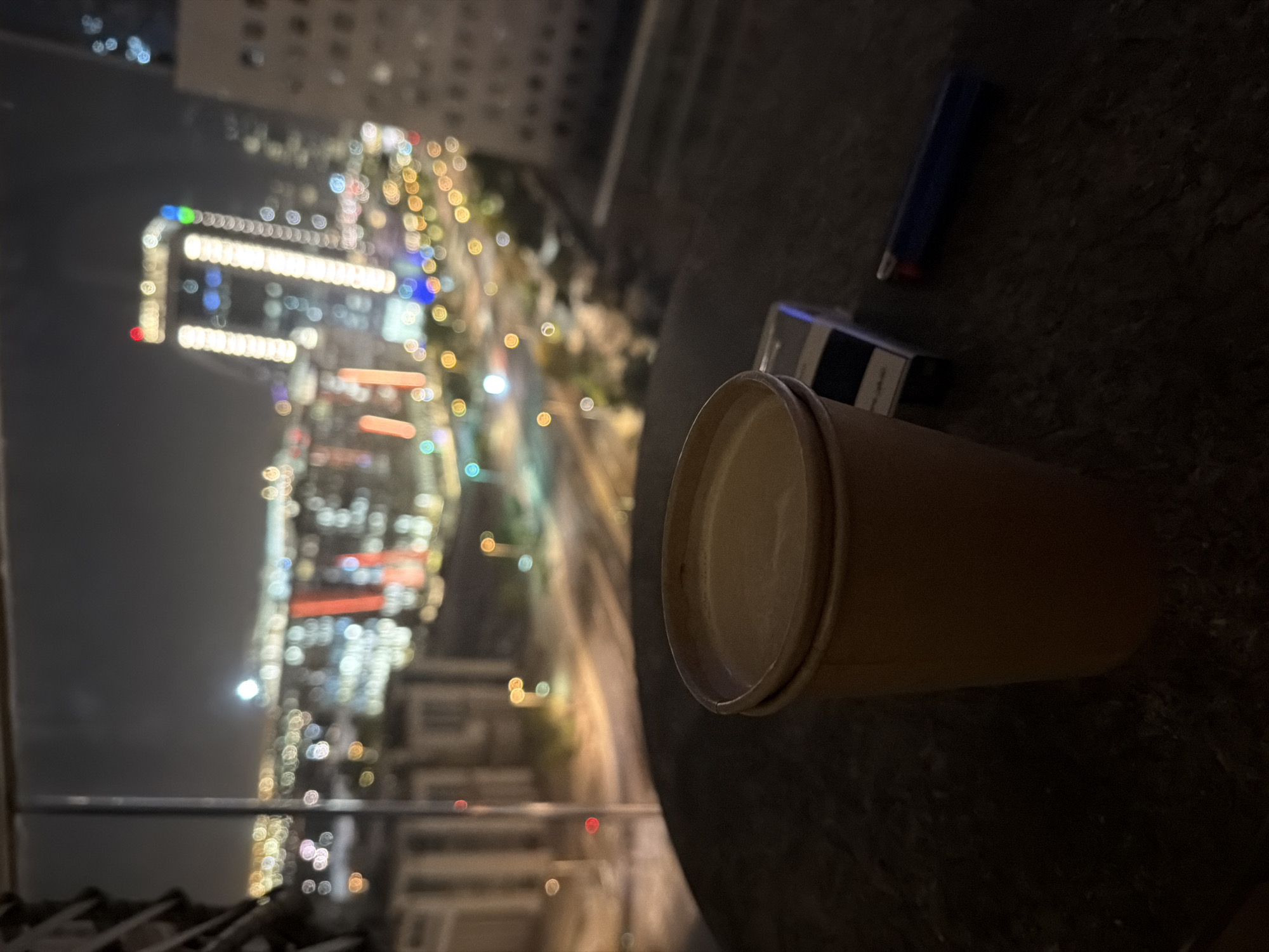 A beverage in a disposable cup is in the foreground of the image, to the left. The cup is light brown and appears to be filled with a light-colored liquid. Next to the cup, to the right, is a pack of cigarettes and a blue lighter. The items are on a dark, textured surface. In the background, a blurred cityscape is visible at night, with numerous illuminated buildings and lights from streetlights and vehicles. The scene appears to be viewed from a high vantage point, possibly a balcony or window.