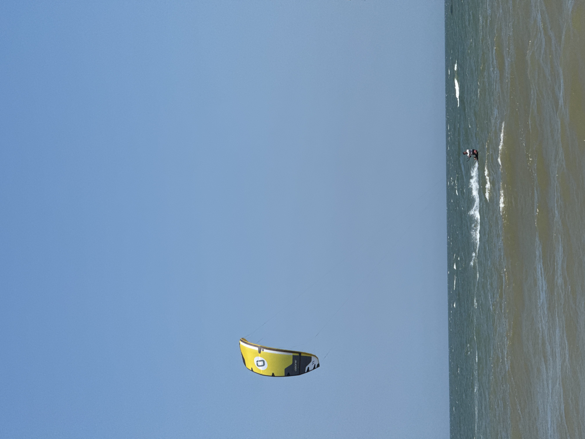 A bright yellow and black kite is visible against a clear blue sky, with the text "OZONE" printed on it. The kite is connected by lines to a person who is kitesurfing on a choppy, greenish-brown ocean. The kitesurfer, wearing a white shirt and dark shorts, is generating a spray of water as they ride. The scene is likely Mũi Né, Vietnam, a popular destination for kitesurfing, known for its strong winds and wide beaches.