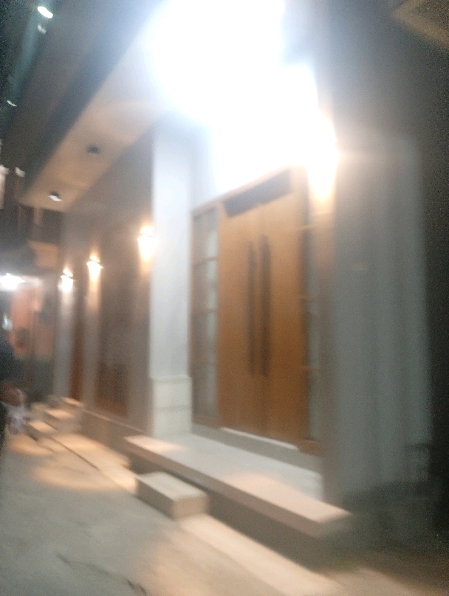 The image captures an exterior view of a building in Banowan, Indonesia. The building appears to be a commercial establishment or a residence, with a wide entrance featuring double doors. Several wall-mounted light fixtures are visible along the exterior wall, casting a warm glow. A pathway or sidewalk leads up to the entrance, with a set of steps providing access. The overall scene is dimly lit, with strong highlights and shadows due to the lighting conditions, and the image quality is poor due to motion blur. No individuals are clearly discernible in the frame.