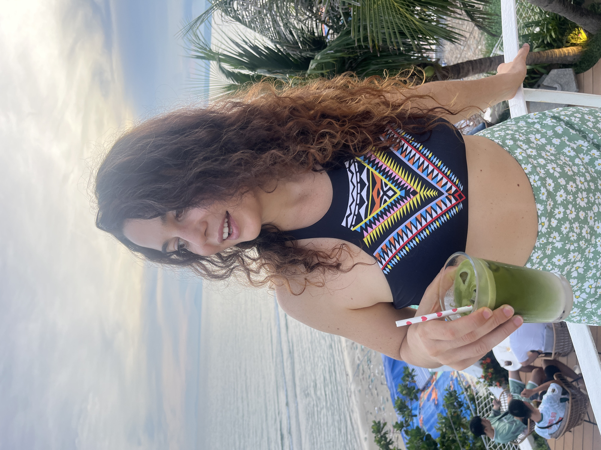 A young woman with curly brown hair stands on a balcony, holding a glass of green matcha tea with a red and white striped straw. She is wearing a black bikini top with a colorful geometric pattern and a light green floral skirt. The background shows the ocean and a beach with palm trees. There are two men sitting on chairs in the lower left corner of the image. The scene appears to be a resort or cafe in Mũi Né, Vietnam, given the presence of palm trees and the ocean view, characteristic of the coastal town known for its beaches and sand dunes. The lighting suggests it might be late afternoon or early evening.