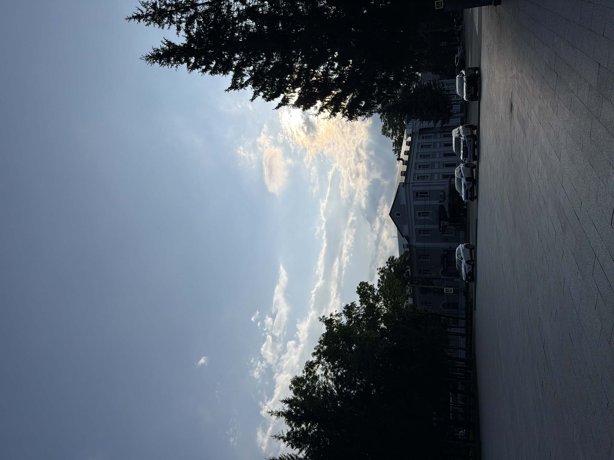 The image depicts an outdoor urban scene in Birobidzhan, Russia, under a partly cloudy sky. In the foreground, a large, paved area with a geometric pattern is visible, appearing to be a public square or plaza. Several dark green, coniferous trees line the middle ground, silhouetted against the sky. Behind the trees, a multi-story building with a light-colored facade and numerous windows is situated. Several cars are parked or driving in the middle ground and background, including a white SUV and a dark-colored sedan. The sky is a mixture of blue and white, with some wispy clouds and areas where the sun is partially obscured, casting a soft light. The overall impression is a quiet urban environment at dusk or dawn.