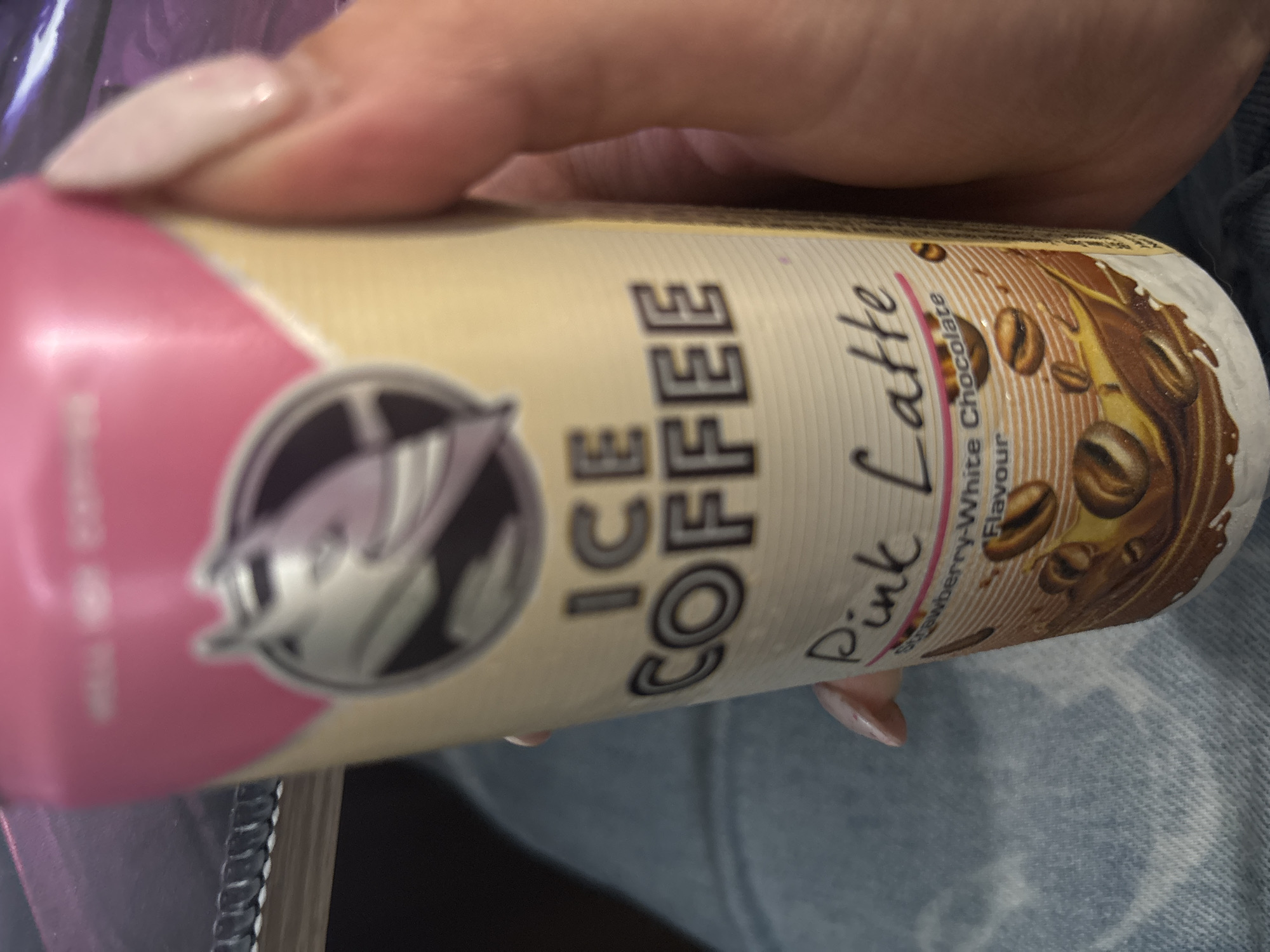 A close-up image shows a person's hand holding a canned beverage, oriented vertically with the can mostly centered. The hand, visible from the knuckles down, has long, light pink or nude-colored acrylic nails.
The can itself is primarily cream or light beige, with a distinct pink section on the left side. Prominently displayed in large, grey, capital letters on the can is the text "ICE COFFEE." Below this, in a pink, handwritten-style font, is "Pink Latte." Further descriptive text, in smaller white letters, reads "Strawberry-White Chocolate Flavour." The can's design also features illustrations of coffee beans and a brown liquid splash, possibly representing coffee or chocolate, on the right side. On the pink portion of the can, there is a circular logo containing a stylized bird or creature.
The background is out of focus, revealing hints of blue denim fabric, suggesting the person might be wearing jeans. The lighting is soft and appears to be indoors.