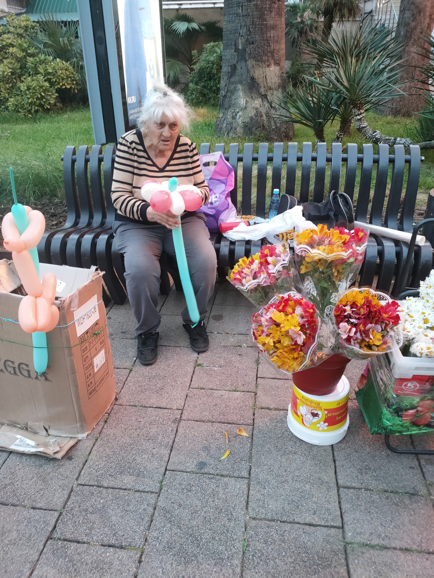 An elderly woman with white hair is sitting on a park bench, holding a balloon animal. She is wearing a striped sweater and grey pants. In front of her, on the ground, are several bouquets of flowers and a cardboard box with balloons. A plastic water bottle and bags are on the bench next to her. The scene appears to be outdoors, with palm trees and greenery visible in the background. This image was taken in Sochi, Russia.