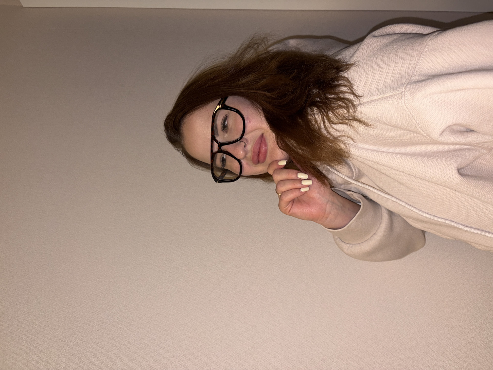 A woman with auburn hair wearing black-rimmed glasses and a light-colored hooded sweatshirt poses for the camera. Her right hand is raised near her chin, with light-colored nail polish visible on her fingernails. The background is a plain light beige wall. The image appears to have been taken indoors under artificial lighting. There is no indication of the location being Krasnodar, Russia.