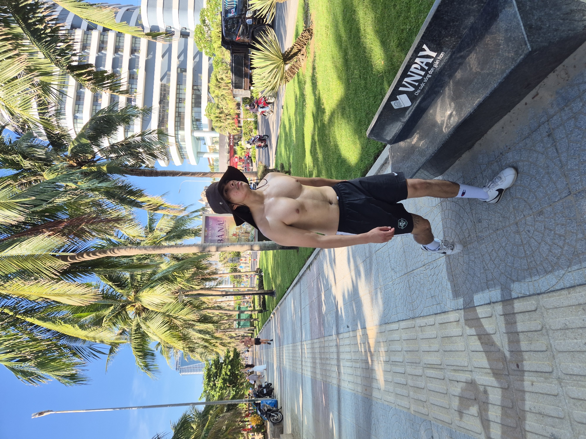 The image captures a sunny day in Da Nang, Vietnam, with a man walking on a paved walkway lined with palm trees. He is shirtless, wearing black shorts, a wide-brimmed hat, white socks, and sneakers. To his right, a modern building with curved balconies stands tall, and a black dump truck is parked on the street. In the foreground, a black bench with the "VNPAY" logo is visible. Further down the walkway, other individuals can be seen walking and cycling, and several motorcycles are parked on the side of the road. The scene suggests a vibrant urban environment near the coast, with lush greenery and clear blue skies.