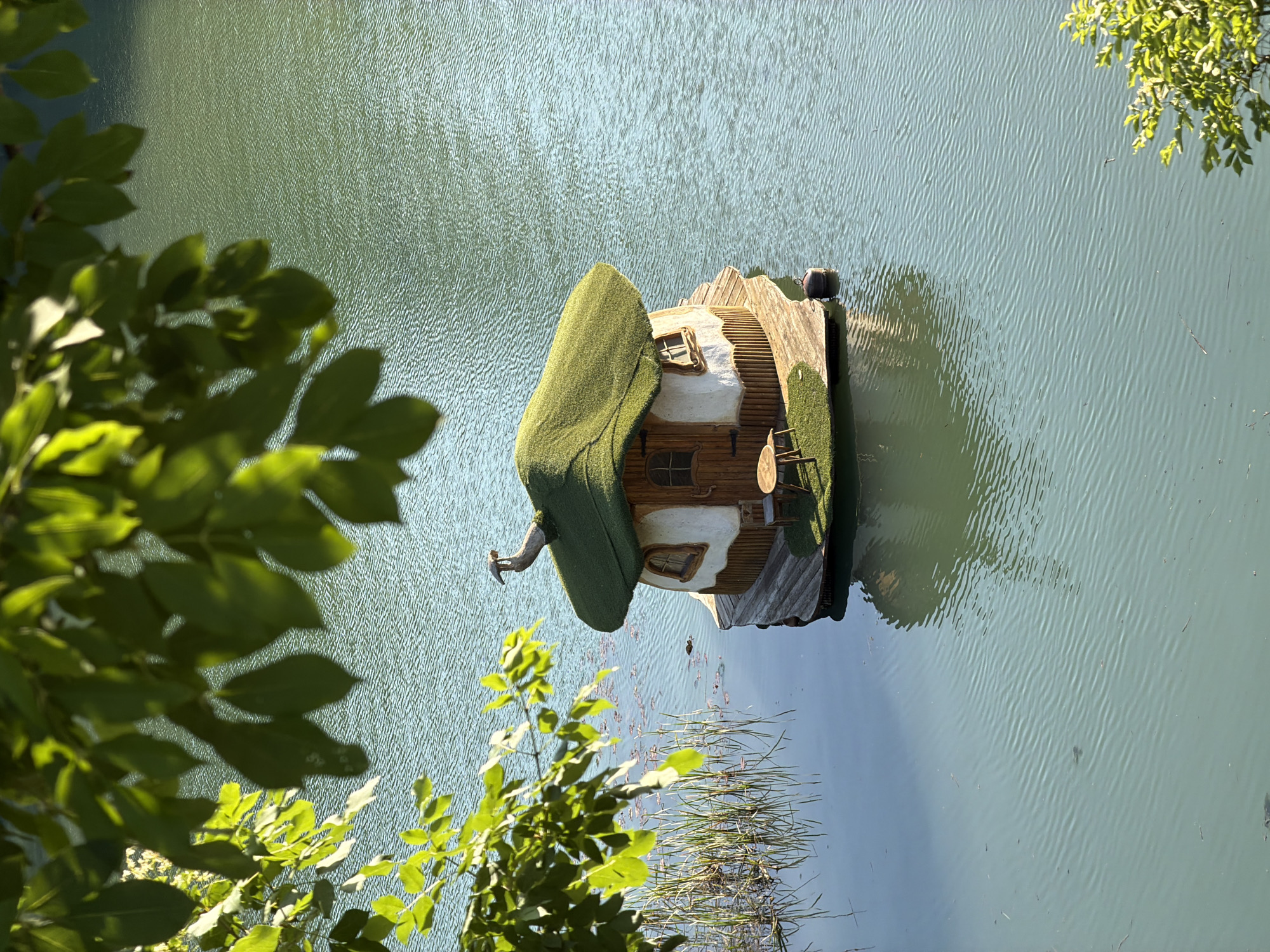 A floating house is located on the water in the city of Lukovit, Bulgaria. The house has a green roof and a chimney. The water is calm and reflects the house and surrounding trees. The house has stairs up to the roof and a small table and chair on its deck. The house is wooden with white accents around the windows and doors. Green shrubbery is next to the back of the house on the deck.