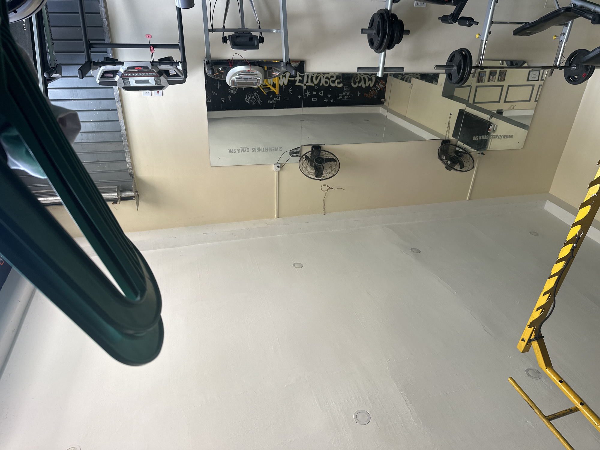 This media file captures an indoor view of a fitness center located in Iddo Tudunwada, Nigeria. The image is taken at an angle and is oriented sideways, showcasing a functional gym space that appears currently unoccupied.
In the center of the background, a large rectangular mirror covers a significant portion of the wall, reflecting elements of the room including other parts of the cream-colored walls, framed pictures, and a black wall with abstract white graffiti. On this mirror, the business name "GIVIEM FITNESS GYM & SPA" is prominently displayed in capital letters, both forward and backward due to reflection. Below the mirror, two black electric fans are visible; one is mounted on the wall, and another smaller one stands on the floor. The graffiti on the wall behind the mirror includes fragmented text and symbols such as "W-FITNESS", "Veige", "Jo", and "D".
To the left, a treadmill with a visible console, screen, and control buttons is positioned against a wall. Above the mirror, a sturdy weight rack holds a barbell loaded with multiple black weight plates, alongside other metallic gym equipment, possibly a bench press or a multi-gym station. The floor is a light, almost white, concrete surface, featuring several circular recessed lights. In the immediate foreground on the left, a dark green, U-shaped plastic object, possibly part of a swing or an exercise aid, partially enters the frame. On the far right side of the image, a bright yellow metal structure, resembling a ladder or support frame with black striped markings, extends from the floor upwards. The room is well-lit, suggesting either ample natural light or strong artificial lighting. No individuals are present in the gym.