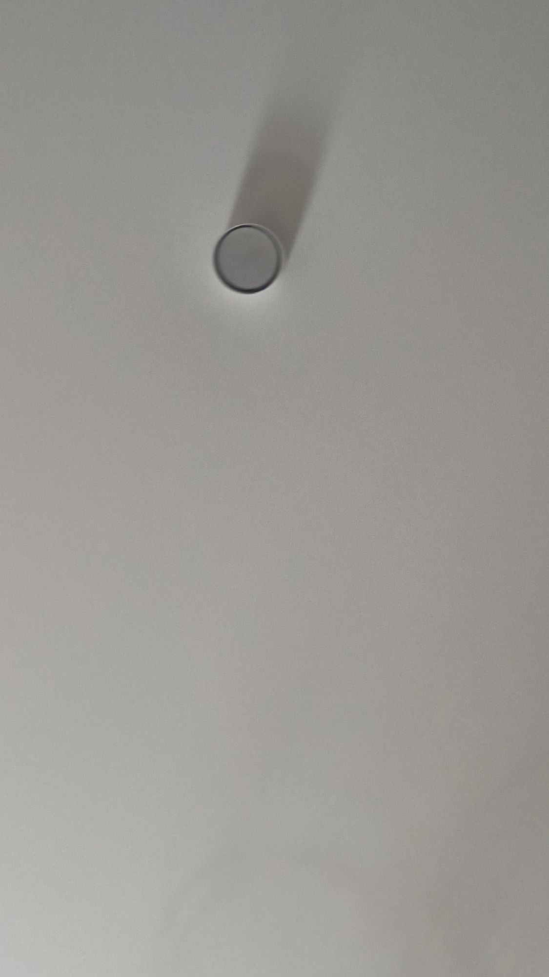 A single, circular object with a metallic sheen is affixed to a plain, light-colored surface, likely a ceiling. The object is casting a long, elongated shadow, indicating a light source from the upper left. The surface appears smooth and is uniformly lit, with no other discernible features or objects present in the frame. The image does not provide any contextual information about the location within the city of Kovrov, Russia, or any details related to people, actions, or interactions.