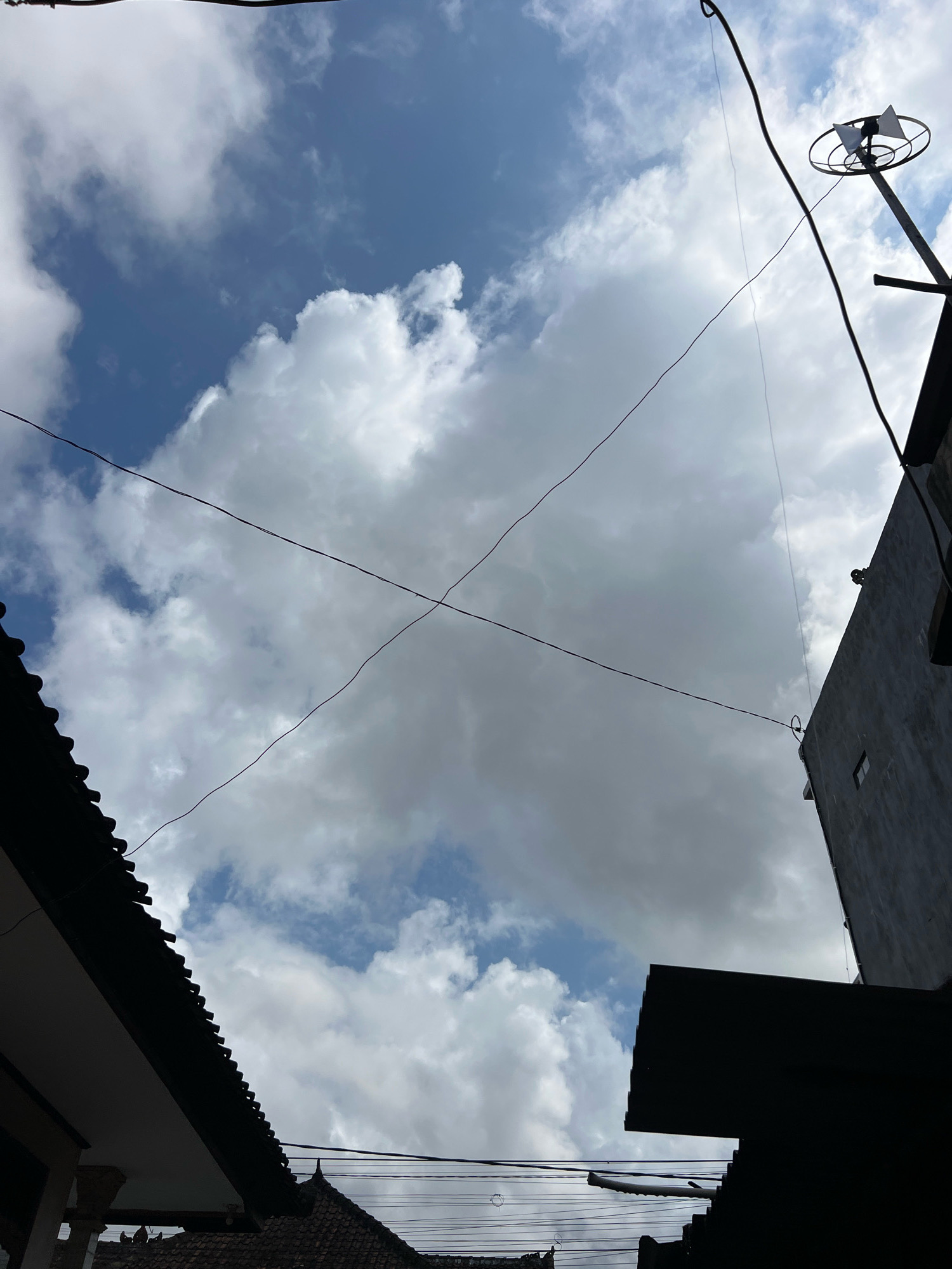 The image is a low-angle shot looking upwards at a cloudy sky in Denpasar, Indonesia. In the foreground, silhouetted against the sky, are the dark outlines of several rooftops, including one with a distinctive tiered pattern and another with a white soffit. Electrical wires crisscross the sky, with one set forming an 'X' shape. To the right, a portion of a grey building is visible, and further up, a metal pole with a circular antenna and two triangular reflectors is mounted. The sky is a mixture of bright blue patches and large, white, fluffy clouds.