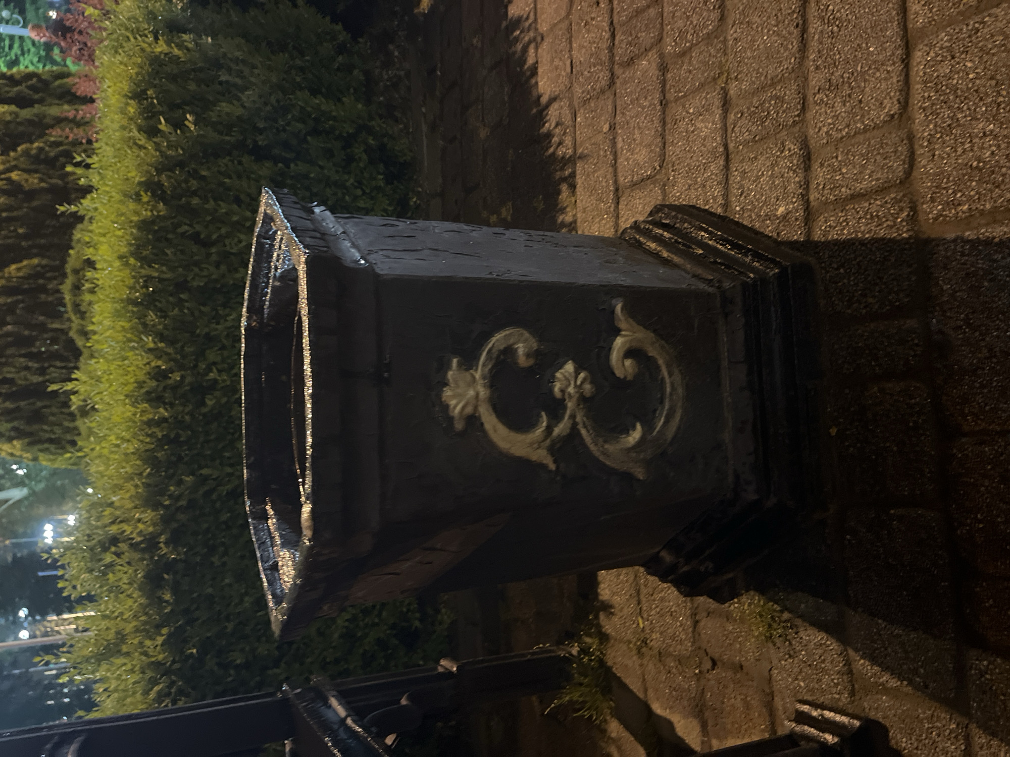 A black, ornate trash receptacle sits near a hedge and a brick walkway. The receptacle is likely located outdoors in a public area. No people are visible in the image. The location of the receptacle may be in the city of Krasnodar, Russia.