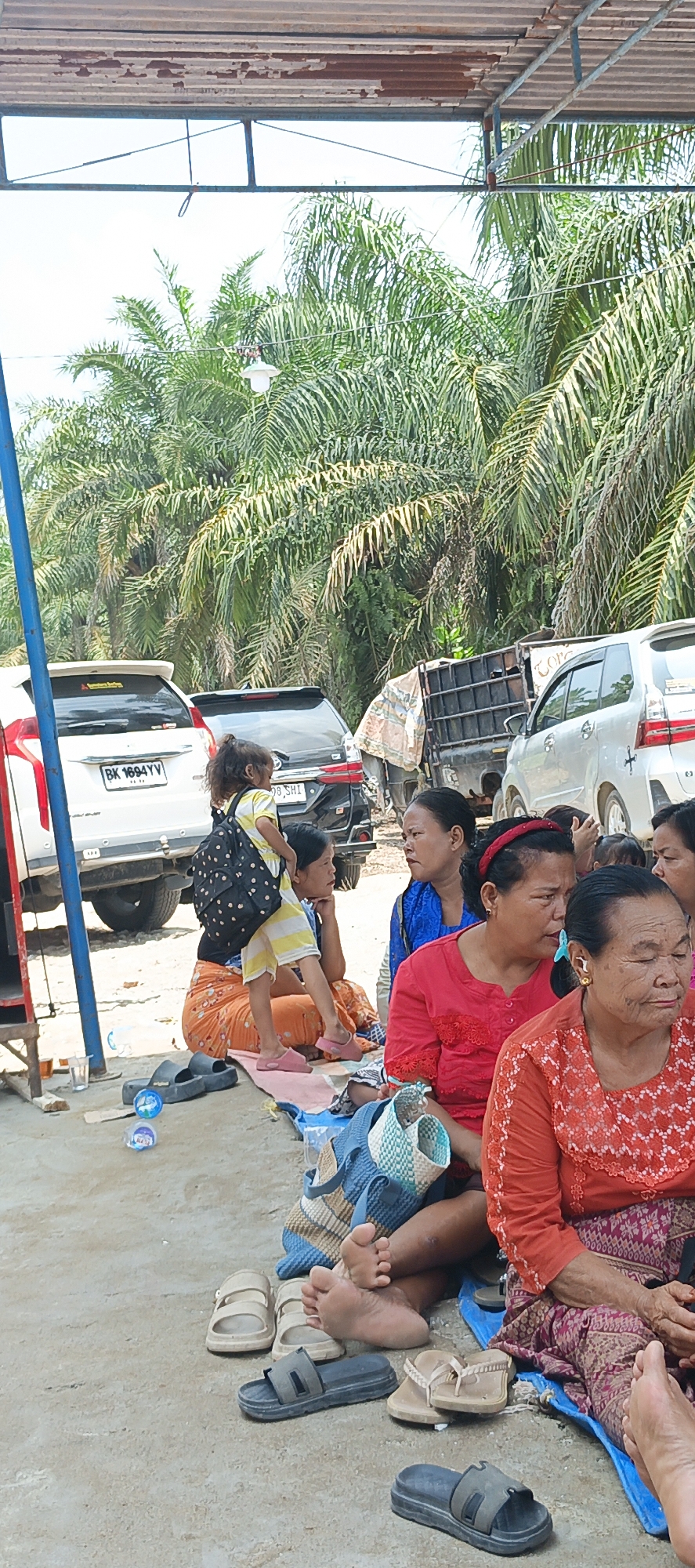 In Negeri Lama, Indonesia, a group of people are gathered outdoors under a makeshift shelter with a corrugated metal roof. Several vehicles, including a white SUV and a black SUV, are parked in the background, suggesting a gathering or event. The ground is a mixture of dirt and sand.
The foreground features several individuals, primarily women, sitting on the ground. A young girl with curly hair, wearing a yellow and white striped shirt and a black backpack, is perched on the shoulders of a woman. The woman is wearing an orange skirt. Other women are dressed in brightly colored clothing, including a woman in a red top and a woman in an orange top with intricate patterns. They are sitting on blue and patterned mats. Various footwear, including sandals and flip-flops, are scattered on the ground.
The environment is characterized by lush palm trees visible behind the shelter and vehicles, indicating a tropical or subtropical setting. The lighting suggests it is daytime, with bright sunlight casting shadows. A blue pole, likely part of the shelter's structure, is visible on the left side of the image. Several bottles of water are also present. The overall scene appears to be a casual gathering in a semi-rural or outdoor setting within Negeri Lama.