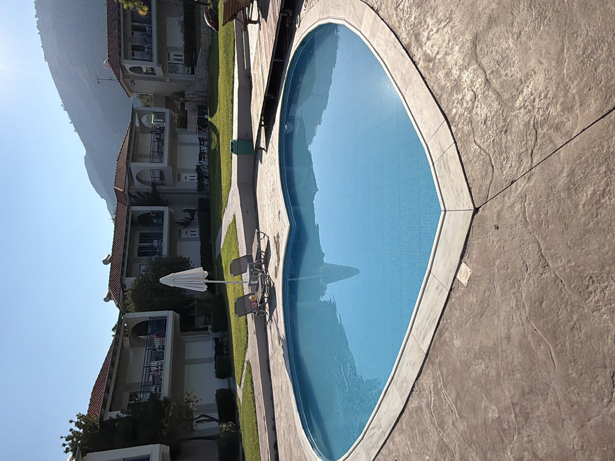 The image depicts a heart-shaped swimming pool filled with bright blue water, surrounded by a textured concrete patio. In the background, there are two buildings with red tile roofs and balconies. A white umbrella stands near the pool, and two lounge chairs are placed on the grass beside it. Further in the background, a lush green lawn leads to the buildings, and behind them, a hazy, mountainous landscape is visible under a clear blue sky. The scene suggests a tranquil resort or residential setting in the Municipal Unit of Alyzia, Greece. There are no people visible in the image.