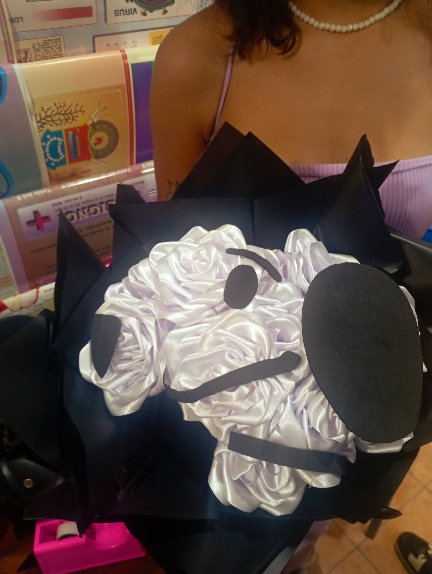 The image shows a close-up view of a person holding a bouquet with a face design. The bouquet features white satin-like fabric arranged to resemble flowers, with black felt pieces forming eyes, a nose, and a mouth to create a character's face. The person, partially visible from the chest up, is wearing a light purple tank top and a pearl necklace. The background consists of shelves with various items, including what appear to be educational posters or charts, some with text in Spanish such as "VIRUS" and "SIGNOS". The scene is indoors, with a tiled floor visible at the bottom right. The image appears to have been taken in Juan Griego, Venezuela, judging by the context provided.