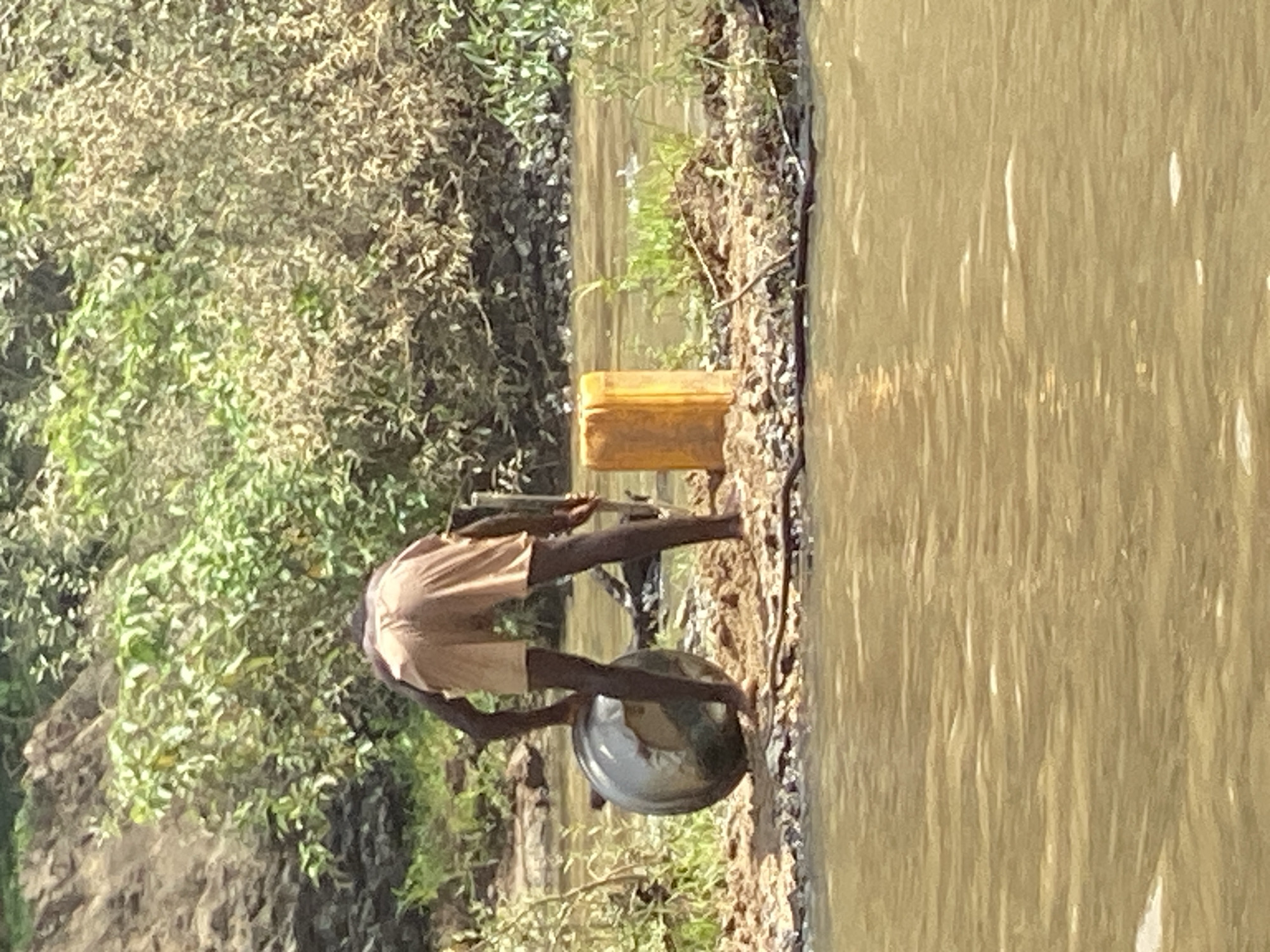 The image depicts an outdoor scene under bright daylight, likely in Kukum, Nigeria. The central subject is an individual, appearing to be an adult with dark skin, wearing light-colored shorts. They are positioned on a dirt or sandy bank next to a body of water.
The person is bent over, seemingly engaged in an activity at the water's edge. They are holding what appears to be a fishing rod or a long stick, suggesting they are fishing or handling fishing equipment. A large, metallic, reflective bowl or pot is situated on the ground near their feet, very close to or partially in the water. Further along the bank, to the left of the person, a rectangular yellow jerrycan or container is visible.
The body of water, likely a river or large pond, is murky and light brown, with reflections on its surface. The bank where the person is standing transitions into dense, green and brown foliage, composed of bushes and trees, which fills the left side of the frame, indicating a natural, perhaps rural or semi-rural, environment. The strong, clear illumination suggests it is daytime, likely sunny. No specific emotions can be discerned, and there is no visible text in the image. The scene captures a moment of practical activity by the water.