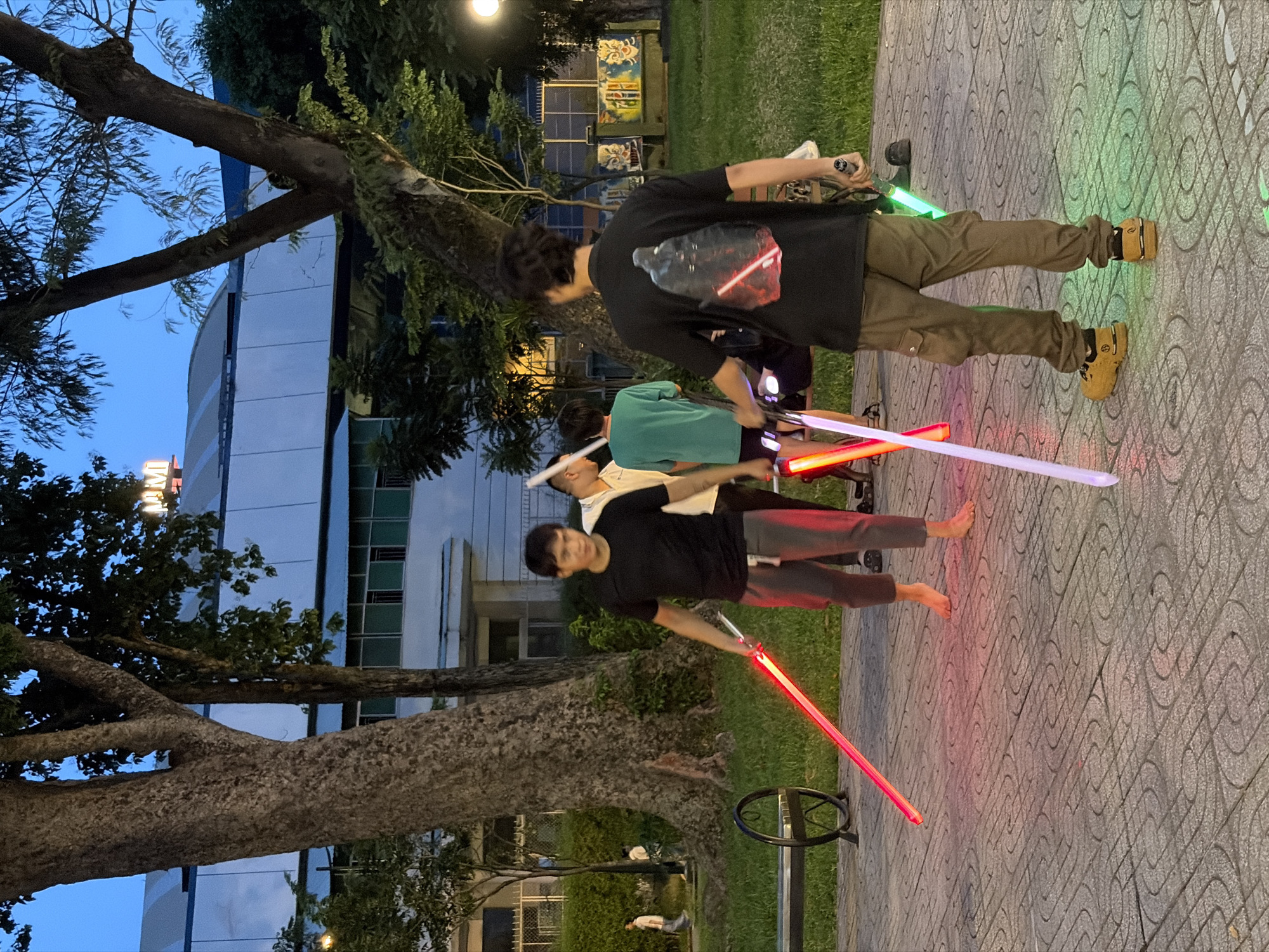 In the city of Thủ Đức, Vietnam, four young men are engaged in an outdoor activity at dusk. Two of them are holding illuminated light sabers, one red and one white, in a paved area with a decorative pattern. Another person, partially visible, also holds a red light saber, while a fourth person stands with a green light saber. They are gathered near a large tree and a park bench, with a building in the background that has "PM" signage. The lighting suggests it is evening, with artificial lights from the sabers illuminating the ground. The overall setting appears to be a public park or urban open space.