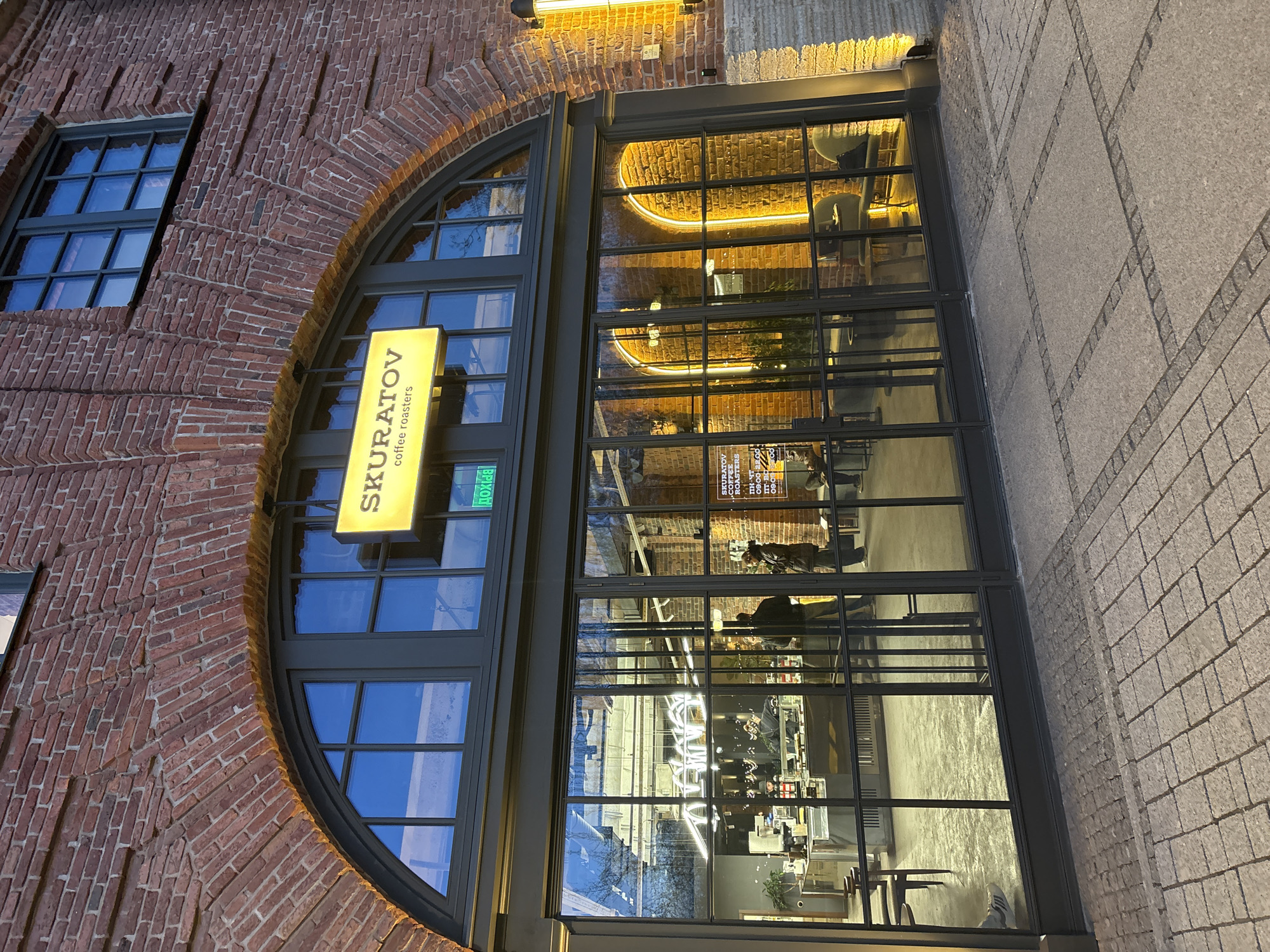 The image captures the façade of a "Skuratov Coffee Roasters" location. The architecture features a prominent brick wall with an arched window arrangement. A rectangular window is visible to the side of the larger arched entrance. The "SKURATOV coffee roasters" sign is positioned vertically.
Inside, the interior space appears to be illuminated, with furniture and patrons visible. Reflections on the glass panes obscure some of the interior details.
The location is determined to be in Saint Petersburg, Russia.
