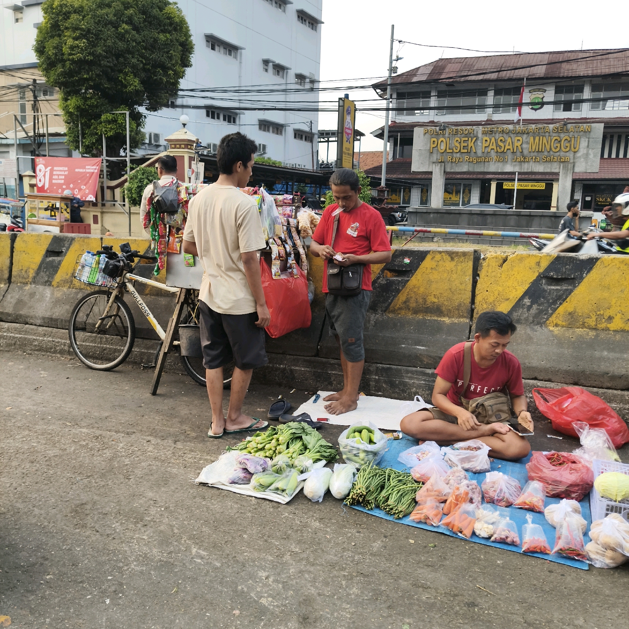 An outdoor street scene depicts a market stall selling produce in front of the POLSEK PASAR MINGGU police station in South Jakarta, Indonesia. A man in a red shirt sits cross-legged on a blue tarp, arranging vegetables and small bags of produce, including chilies, carrots, and onions. Beside him are red plastic bags and a white crate with yellow cabbage. To his left, another man in a red shirt stands, holding money and facing a stall with a bicycle attached. Two other men stand near the stall, one in a tan shirt and dark shorts, the other partially visible in the background. The stall displays a variety of packaged goods and snacks, with a bicycle parked beside it, its basket filled with bottles and cleaning supplies. Yellow and black striped concrete barriers line the background, separating the street from the police station building, which has a prominent sign that reads "POLSEK PASAR MINGGU" and lists the address. The ground is paved with asphalt, with some debris scattered around. The overall atmosphere suggests a busy, everyday street market setting.