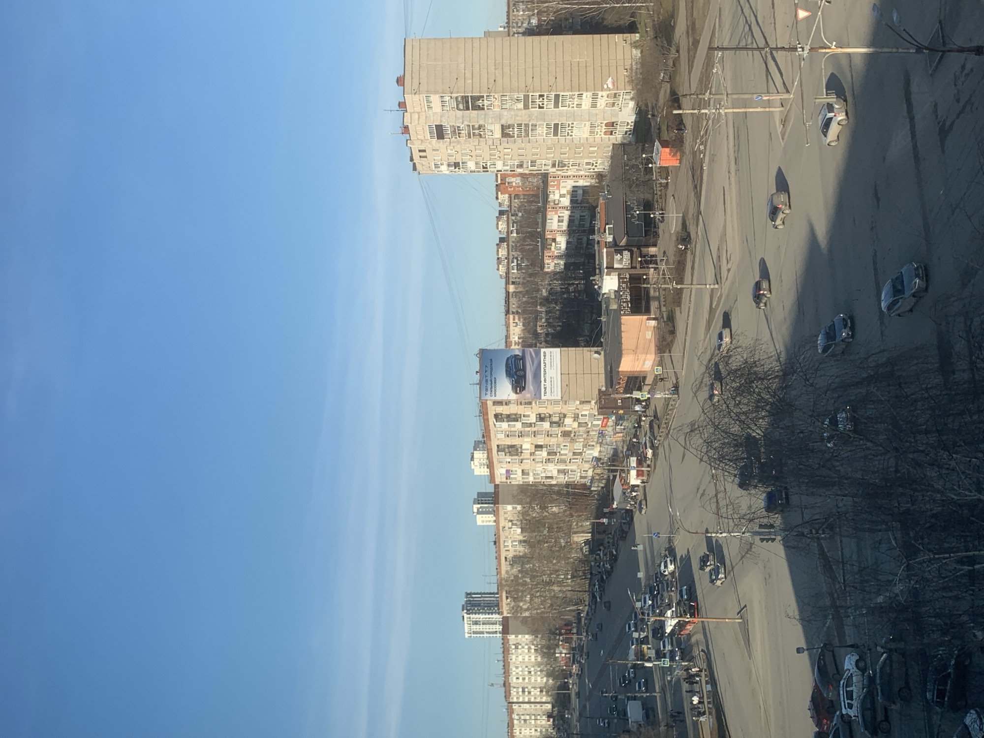 The image shows a cityscape in Izhevsk, Russia, under a clear blue sky. Tall apartment buildings line a wide street bustling with vehicular traffic. The street is divided by a median strip, and trees without leaves are visible along the roadside. A billboard advertising "TENET INTERPARTNER" is prominent among the buildings. Cars of various colors are moving along the street. Overhead, electrical cables stretch between buildings and poles. The scene is taken from an elevated perspective, possibly from a higher building.