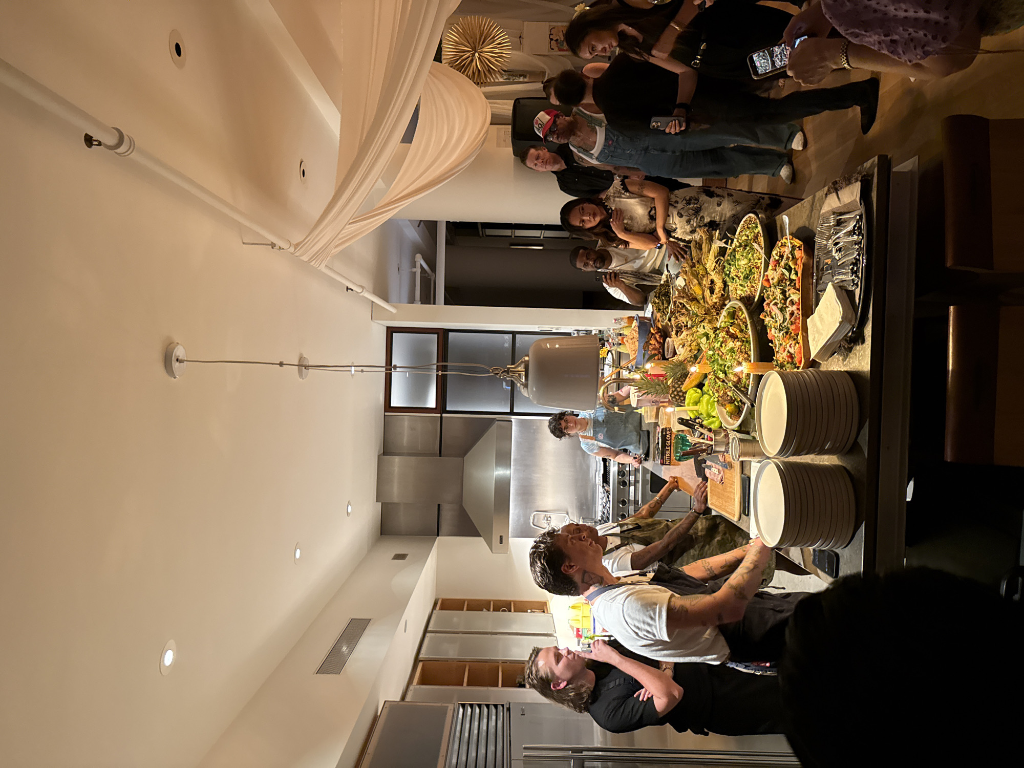 A kitchen scene in New York City, United States of America shows a gathering of people near a long table laden with food. On the left, kitchen staff are preparing food, while a group of guests on the right admire and take pictures of the spread. The table includes multiple salad bowls, plates, and serving utensils. The kitchen itself features stainless steel appliances and cabinetry. Above, decorative lighting and white fabric drape from the ceiling.