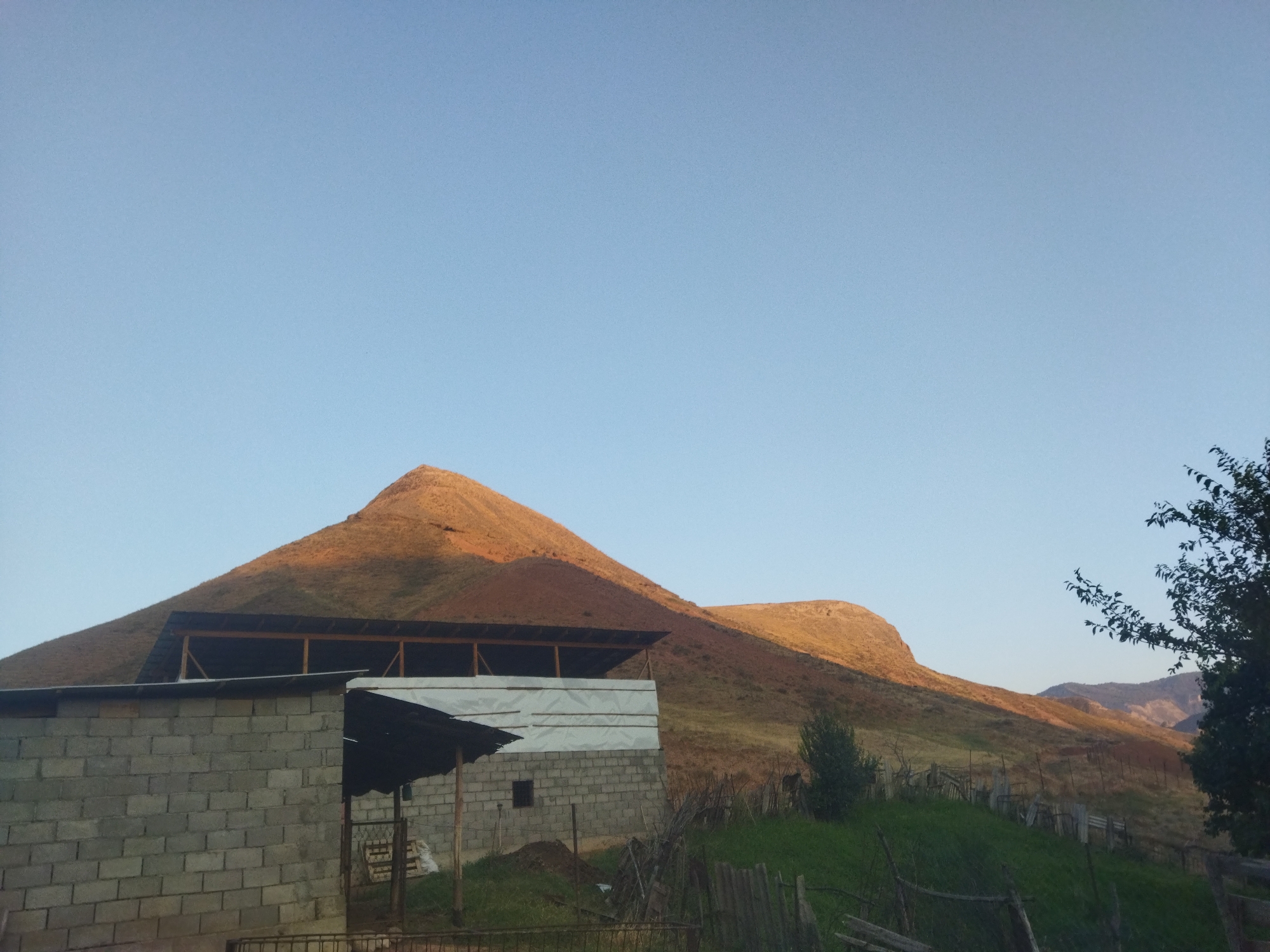 The image depicts a rural landscape in the city of Mailuu-Suu, Kyrgyzstan, during what appears to be sunset or sunrise, given the warm lighting on the hills. In the foreground, there are two unfinished or partially constructed brick buildings. The building on the left is made of grey concrete blocks and has a corrugated metal roof. Adjacent to it, another structure, also of concrete blocks, is partially covered with white sheeting, suggesting ongoing construction or renovation. A wooden fence runs along the bottom of the image, bordering the property. Behind the buildings, rolling hills dominate the scene, with the larger hill to the left bathed in golden light. The hills are covered in sparse vegetation. A tree with dark leaves is visible on the right side of the frame. The sky is a clear, light blue. There are no people visible in the image, and no animals are clearly discernible. The environment appears to be agricultural or semi-rural, with evidence of human habitation and development amidst natural terrain.