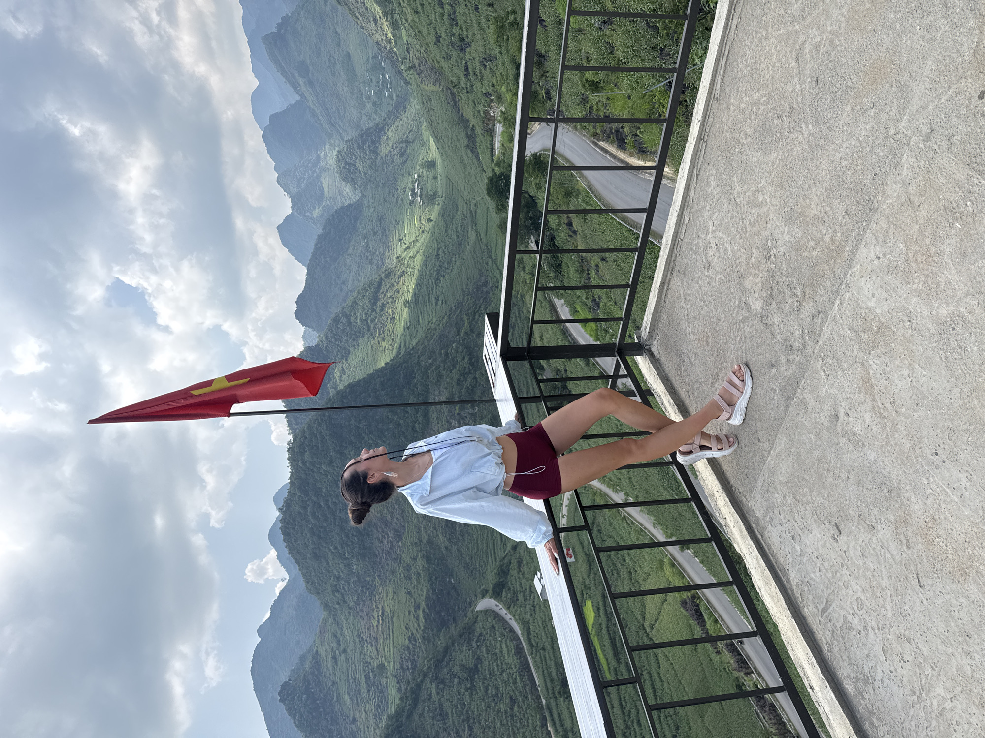 A woman is leaning on a white guardrail, with a mountain range as the backdrop. The location is the Lũng Phìn Commune, Vietnam. She is wearing maroon shorts, a light-colored button-down shirt, and white sandals. A red flag with a yellow star is nearby. A road winds through the green mountainous terrain.