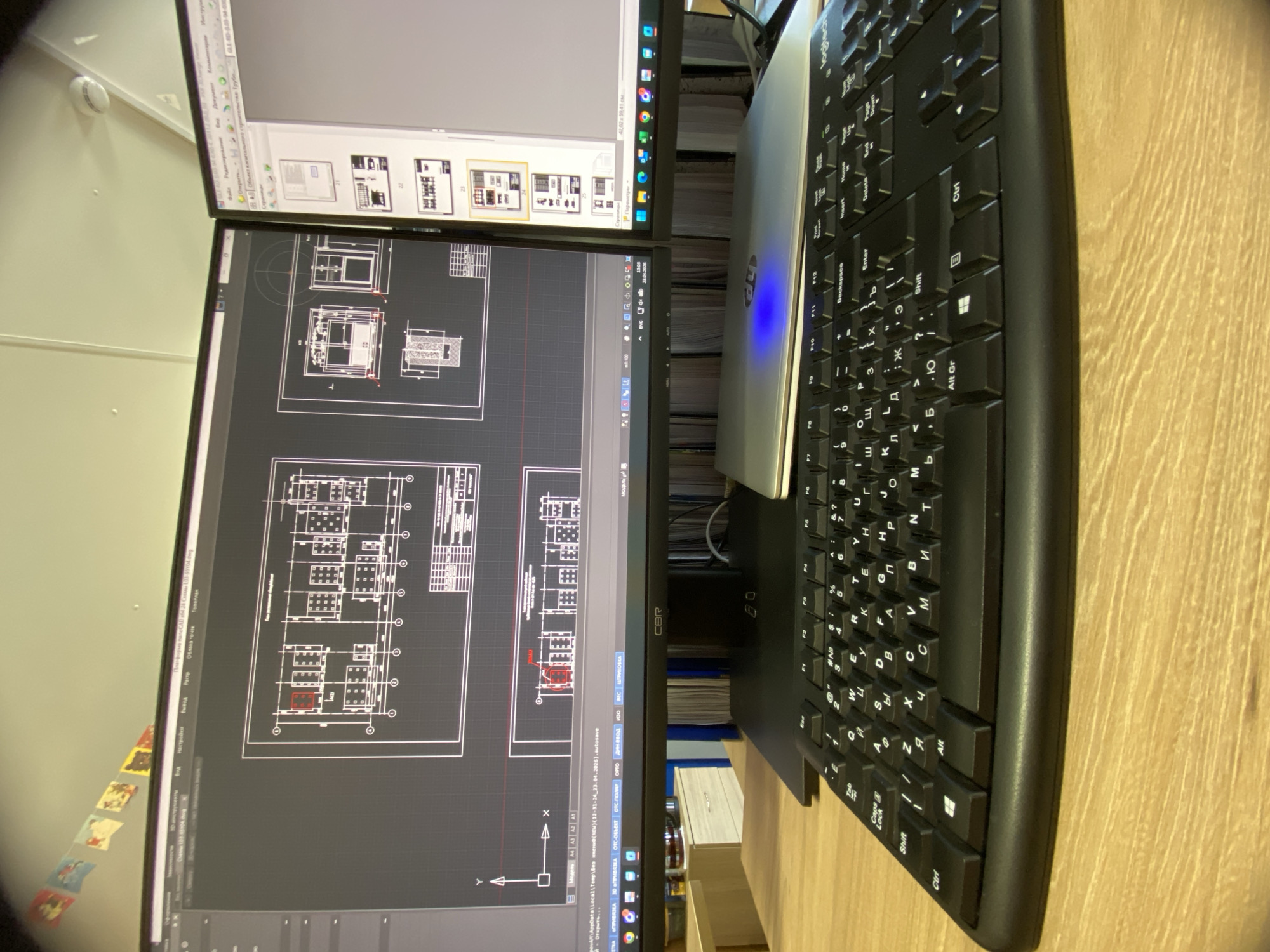 The image shows a workspace with two computer monitors, a laptop, and a keyboard on a wooden desk. The lower monitor displays what appears to be a technical drawing, possibly a floor plan, with lines, shapes, and text annotations. The upper monitor shows thumbnails of images and file names in Russian. A silver laptop is positioned between the monitors with its lid open, partially obscuring the screen. A black keyboard with Cyrillic characters lies to the right, close to the edge of the desk. Based on the software interface shown in the picture, the location depicted is likely associated with architectural or engineering work involving technical drawings and document management. The location of Luzhitsy, Russia cannot be confirmed from the contents of the image.