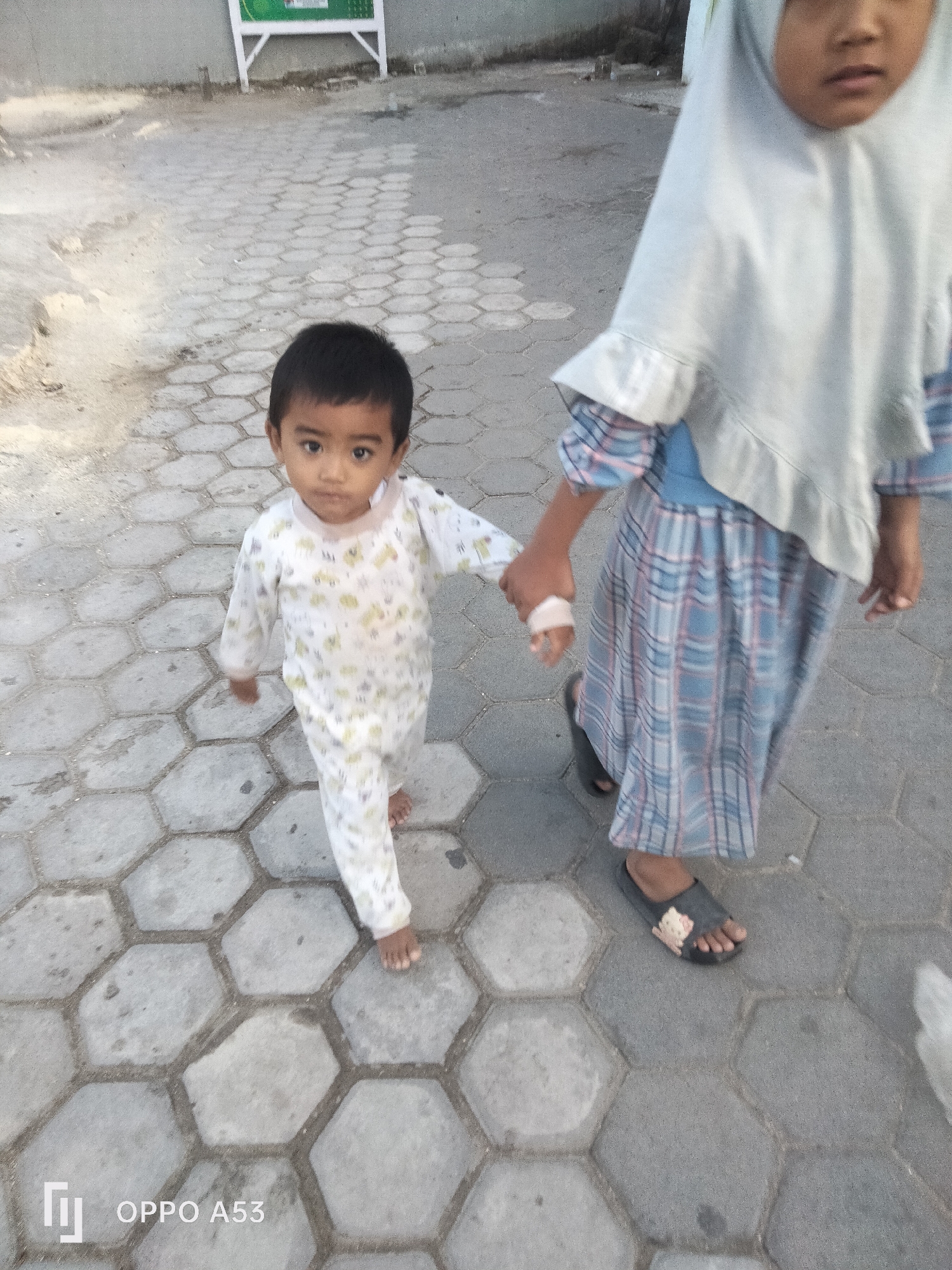 Two children are walking on a hexagonal paved walkway. The child in the foreground, on the left, is a young boy wearing a white long-sleeved pajama set with a yellow floral pattern. He is looking at the camera and holding hands with the child beside him. The child on the right is older and appears to be a girl wearing a light blue hijab and a long blue and pink checkered dress. She is holding a white object in her hand, which has a bandage on one finger. The ground is paved with hexagonal gray stones with sandy grout lines. In the background, there is a concrete wall and a white structure with green signage. The scene appears to be in an outdoor urban environment.