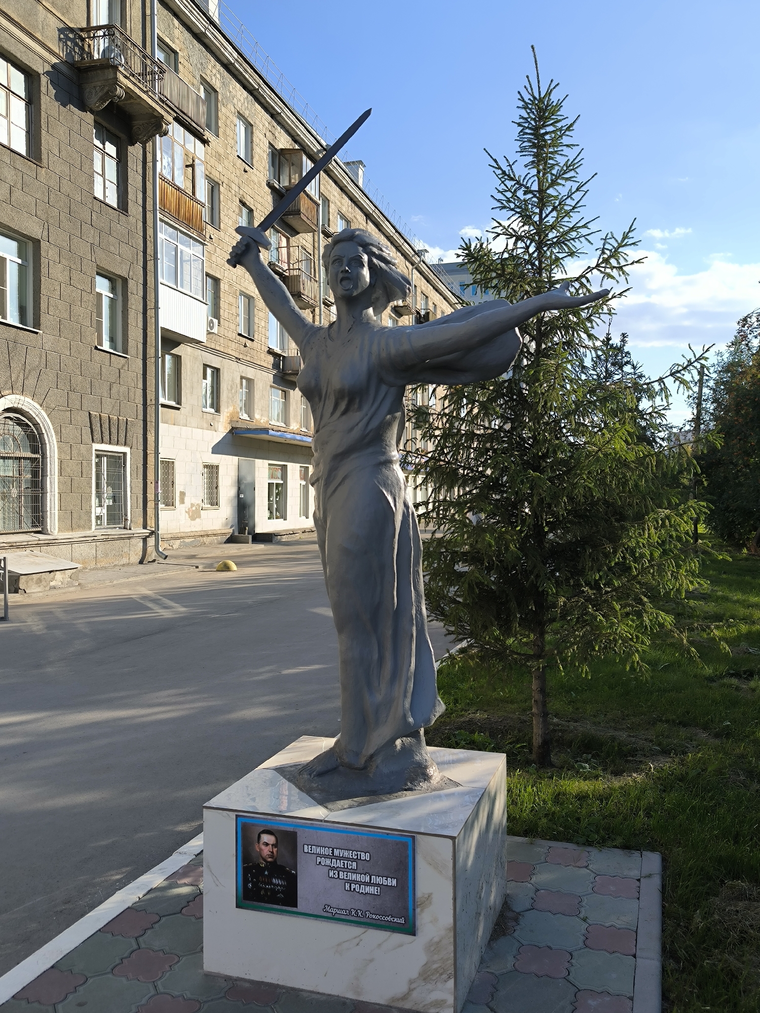 The image shows a statue of a woman with a sword raised in her right hand, her left arm extended. The statue is made of a gray material and stands on a multi-sided pedestal. On the front of the pedestal, there is a plaque with a portrait of a man in military uniform and text in Russian. The man in the portrait is identified as Marshal K.K. Rokossovsky, and the text reads "Great courage is born from great love for the Motherland!". The statue is located on a paved area, with a building in the background. There is a small evergreen tree to the right of the statue, and grass and more trees in the background. The sky is blue with some white clouds. This memorial is located in Novosibirsk, Russia.