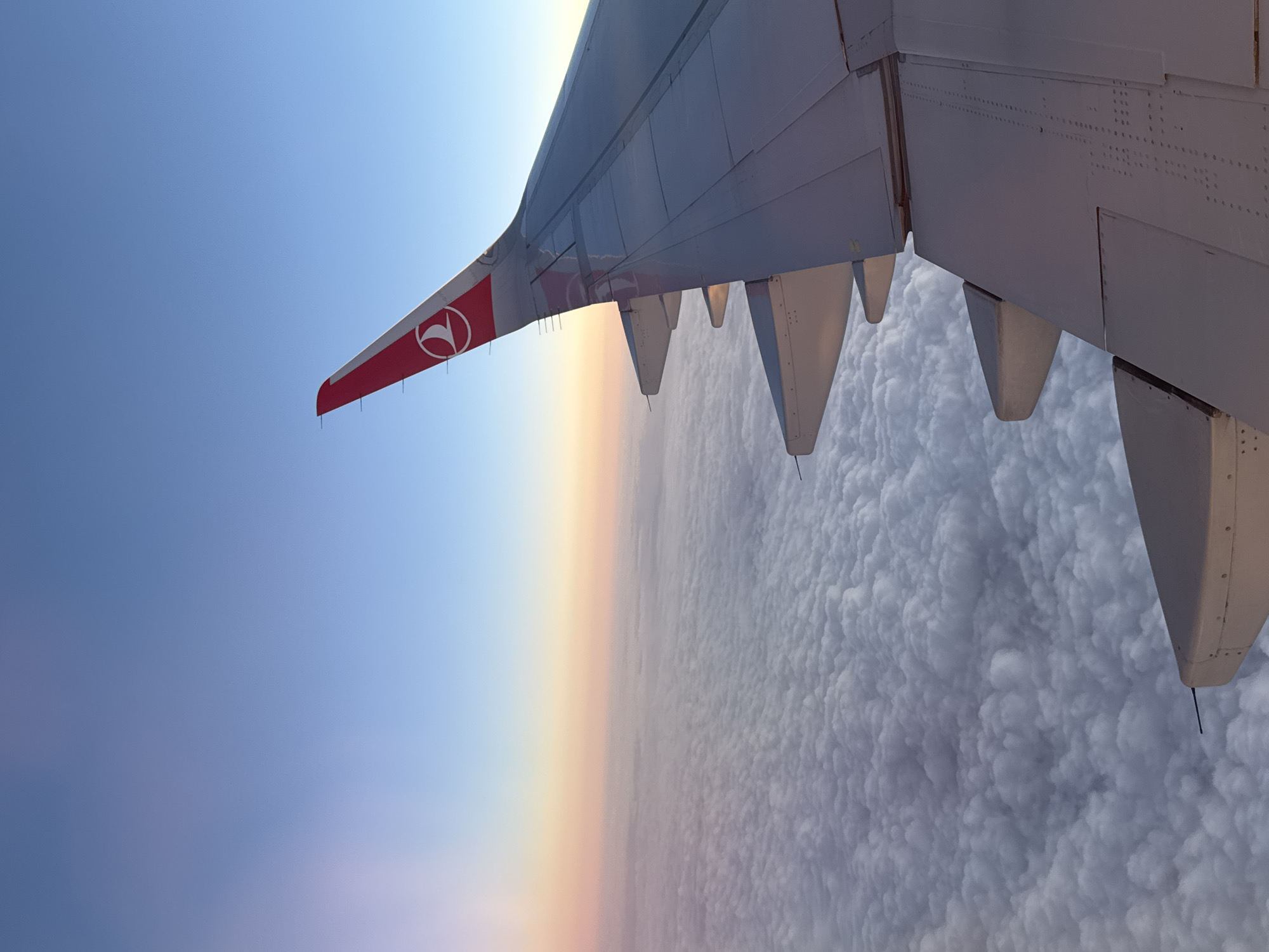The image depicts the wing of an aircraft, with the Turkish Airlines logo visible on the winglet. The sky transitions from blue to a yellowish-orange horizon line, with a dense cloud layer visible below the wing. The location is not determinable.
