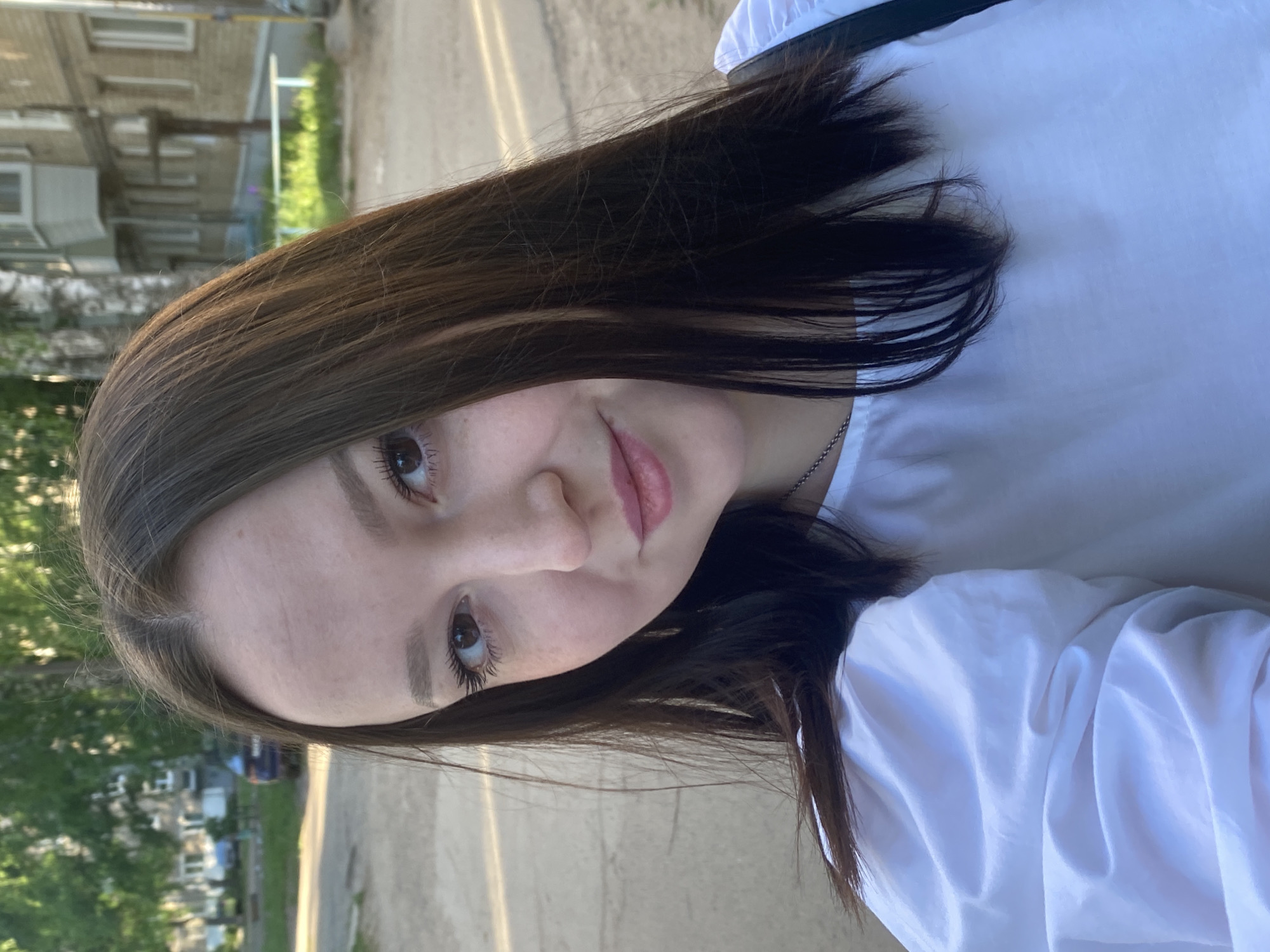 A young woman with dark brown hair is centered in the shot, wearing a white blouse. She is looking directly at the camera with a slight smile.
The background shows a street in what is likely a residential area. Buildings are visible on the left with trees and greenery in front of them. The street appears to be paved with an asphalt-like surface. The time of day appears to be daylight hours.