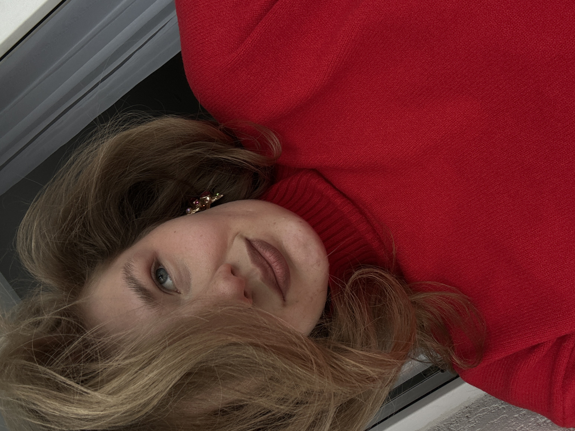 A young woman with light brown hair and fair skin is lying down, facing the camera at an oblique angle. She is wearing a red turtleneck sweater and an earring with multi-colored stones is visible in her left ear. She has blue eyes and light brown lipstick. The background is out of focus, but includes a white and gray surface. The image does not appear to be set in the city of Kazan, Russia.
