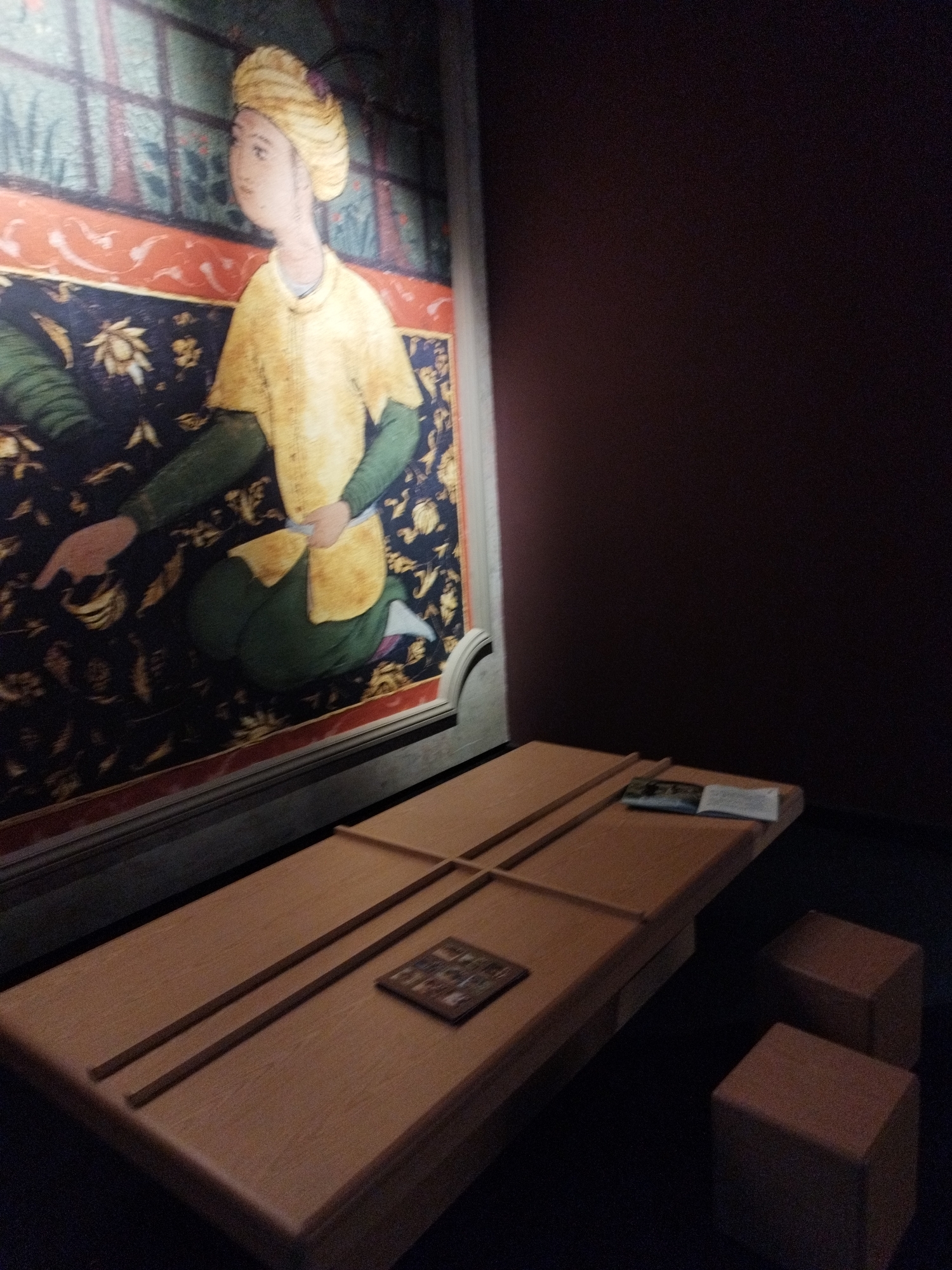 The image depicts an indoor scene featuring a painting and a wooden display table with two small square stools. The painting, which dominates the left side of the frame, is a detailed mural or tapestry showing a person adorned in traditional attire, including a turban and a yellow tunic over green garments. The person is depicted kneeling and pointing. The background of the painting features intricate patterns and floral motifs. To the right of the painting, a long, low wooden table is present. On this table, there are two thin, dark wooden slats running parallel to each other. In the center of the table, a small, framed object, possibly a plaque or a miniature artwork, is placed. Two brown square stools are positioned to the right of the table. The overall environment appears to be a museum or exhibition space, with dark walls and dim lighting. The location is not specified in the image.
