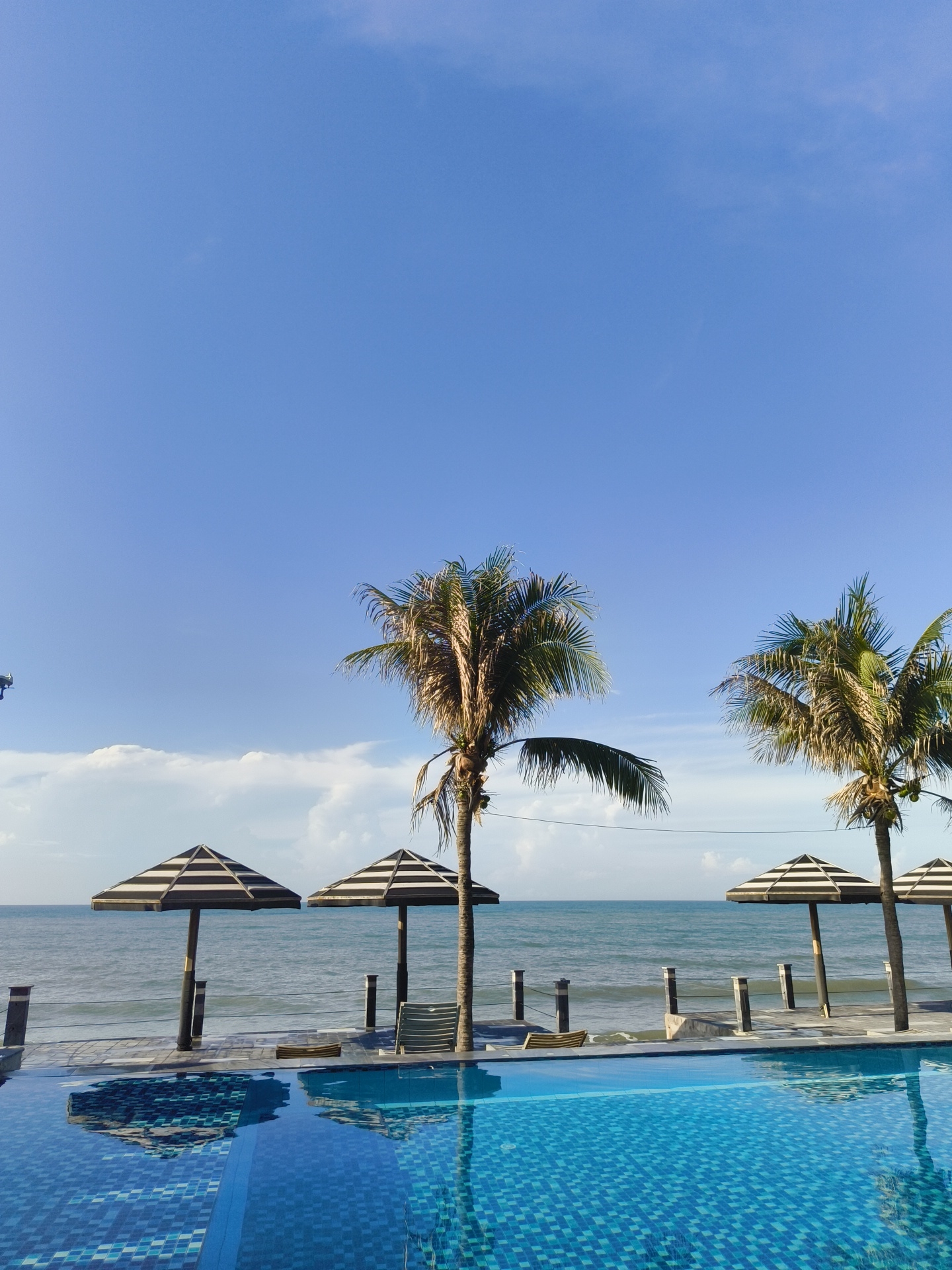 The image depicts a serene coastal scene in Phu Thuy Ward, Vietnam. In the foreground, a pristine blue swimming pool with a tiled bottom reflects the bright sky and surrounding elements. Beyond the pool, a paved walkway lined with railings separates it from the ocean. Three striped umbrellas, alternating black and white, are positioned along the walkway, providing shade. Two prominent palm trees stand between the umbrellas and the ocean, their fronds swaying gently. The ocean stretches out to the horizon, meeting a vast expanse of clear blue sky dotted with a few wispy white clouds. The overall atmosphere is one of tranquility and tropical relaxation.